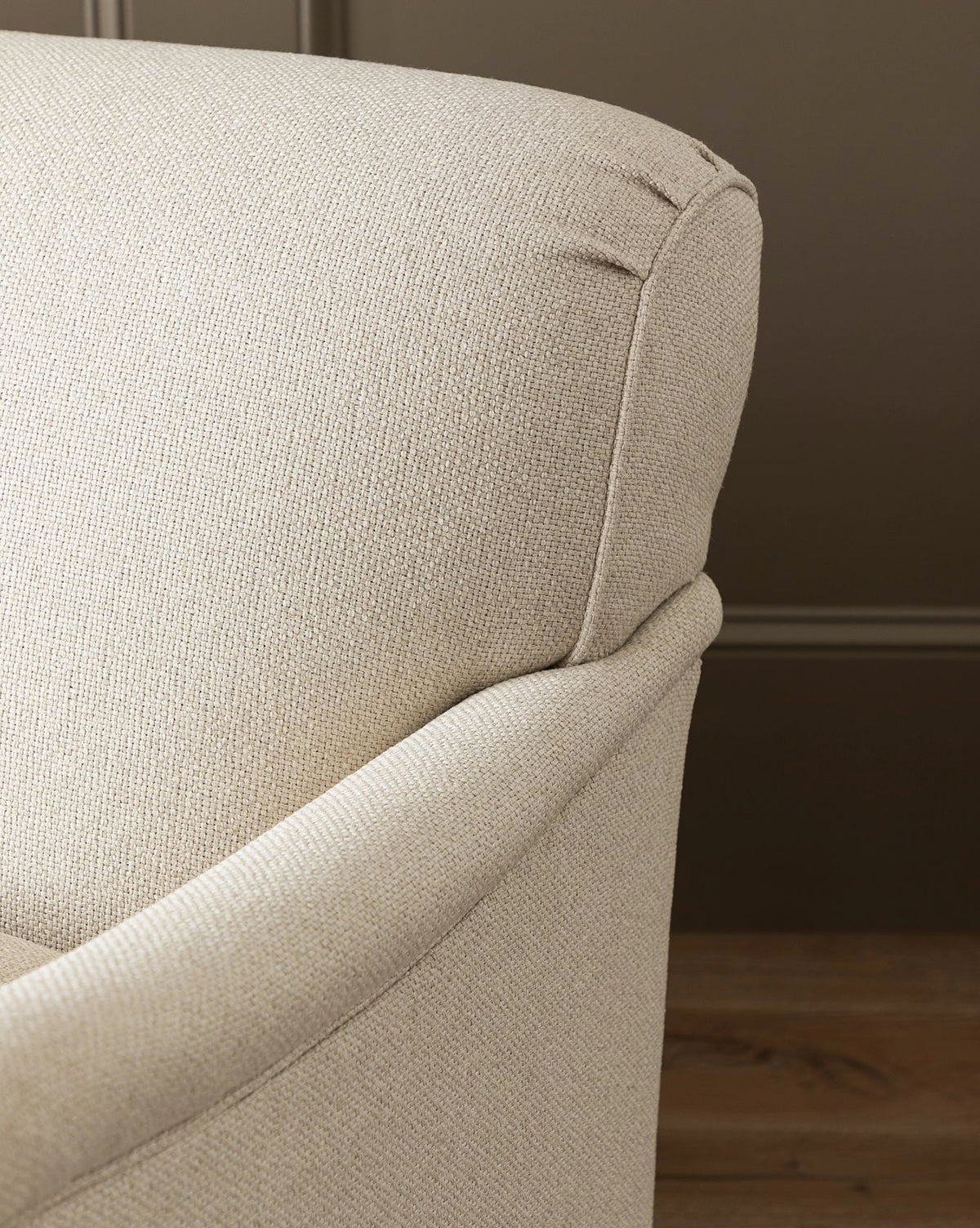 Close-up of the upper corner of the light beige Rowley Sofa 85 by Rowe Fine Furniture, Inc., featuring textured fabric and a curved armrest against a brown wall and wooden floor. Explore customizable fabric options for this hand-crafted piece. Vendixs