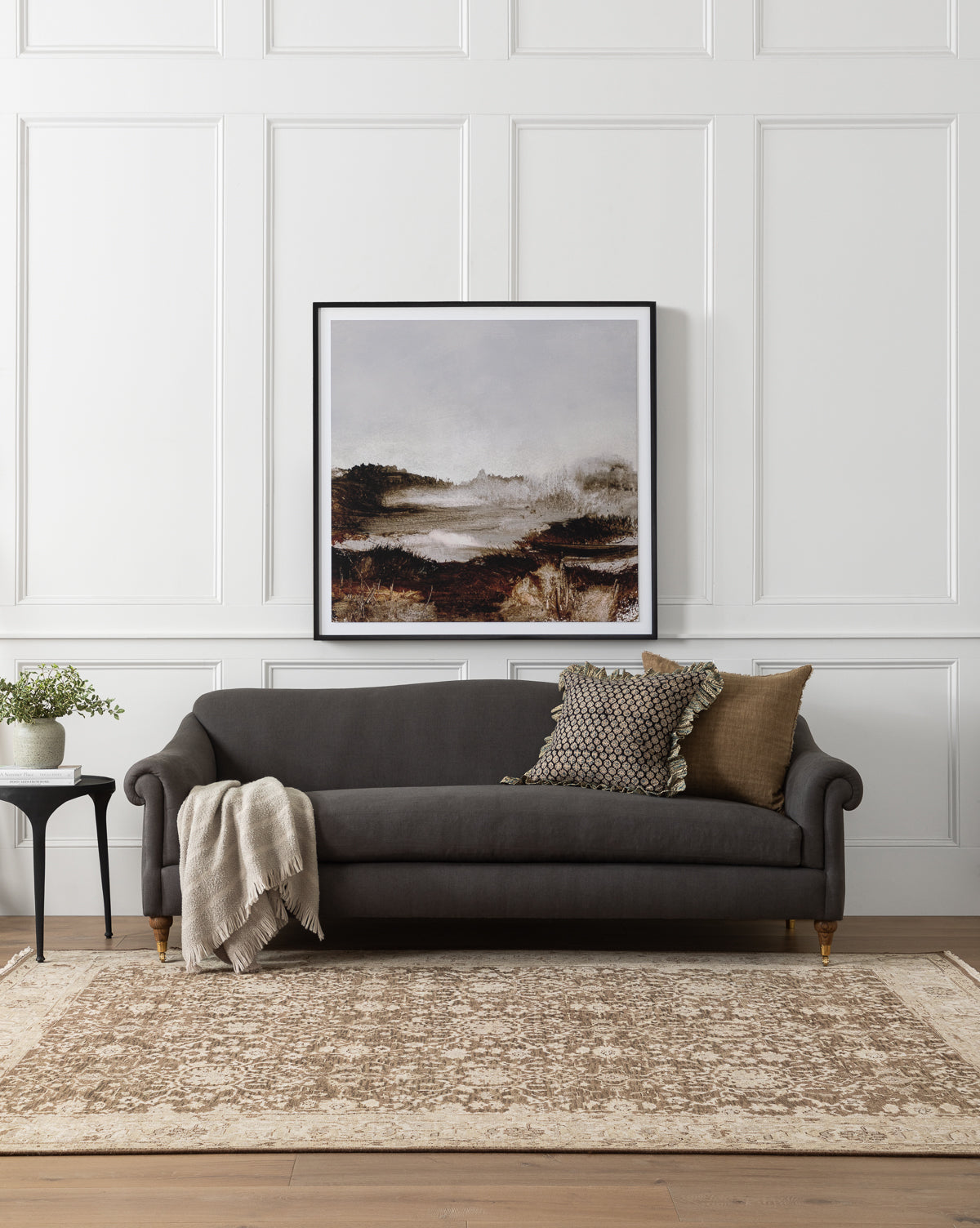A modern living room exudes luxurious comfort with the Community Hildegard Sofa 90 in dark gray, assorted pillows, a soft blanket, side table with a potted plant, beige patterned rug, and a large framed landscape painting on white paneled walls by Vendixs