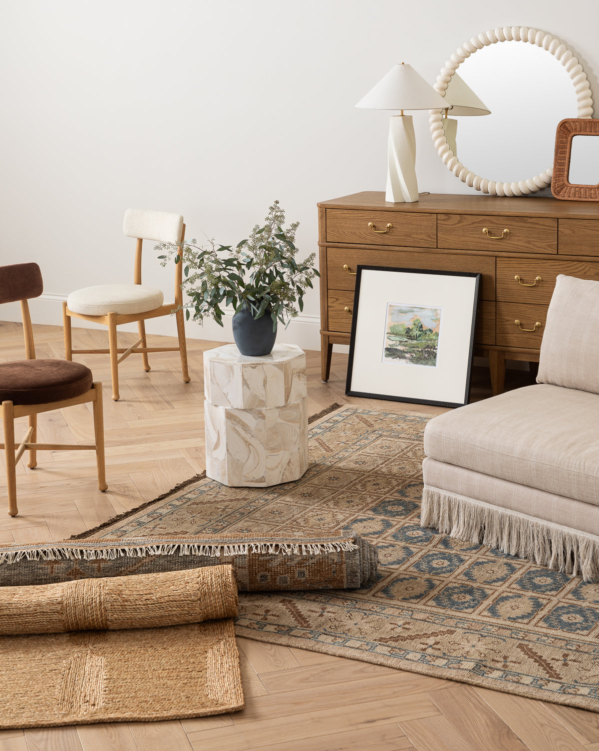 A cozy living room features a wooden dresser with a round mirror, two chairs, a beige fringed lounge seat, the Nunez Hand-Knotted Wool Rug, a small white side table with a plant, and framed artwork leaning against the wall, by Vendixs