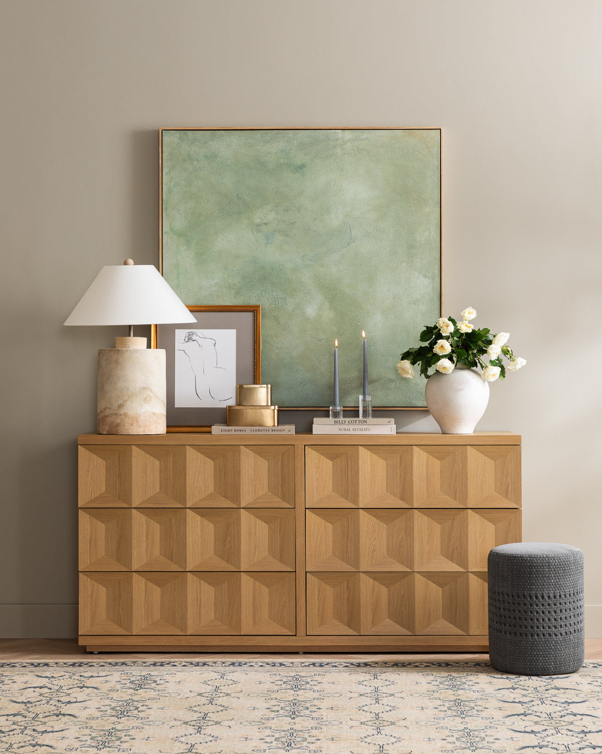 A geometric wooden sideboard is topped with a Samala Lamp, framed art, candles, a white vase of flowers, and vintage accents. Above hangs a large green abstract painting on a neutral wall, with a gray pouf placed close by, by Vendixs