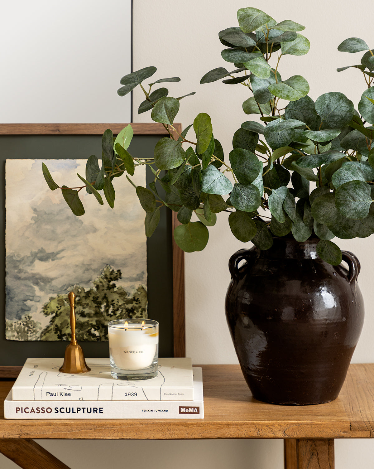 A dark ceramic vase with eucalyptus branches sits on a wooden table next to the Elsher Bell, a candle, art books, and a landscape painting propped against the wall for timeless vintage decor, by Vendixs