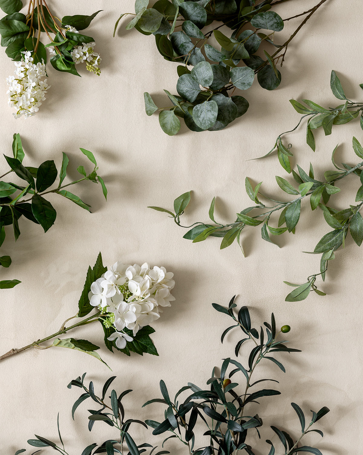 The Faux Gardenia Leaf Stem with green foliage and clusters of small white flowers is artfully arranged on a light beige background, spaced apart to create a natural, minimalist flat lay composition, by Vendixs