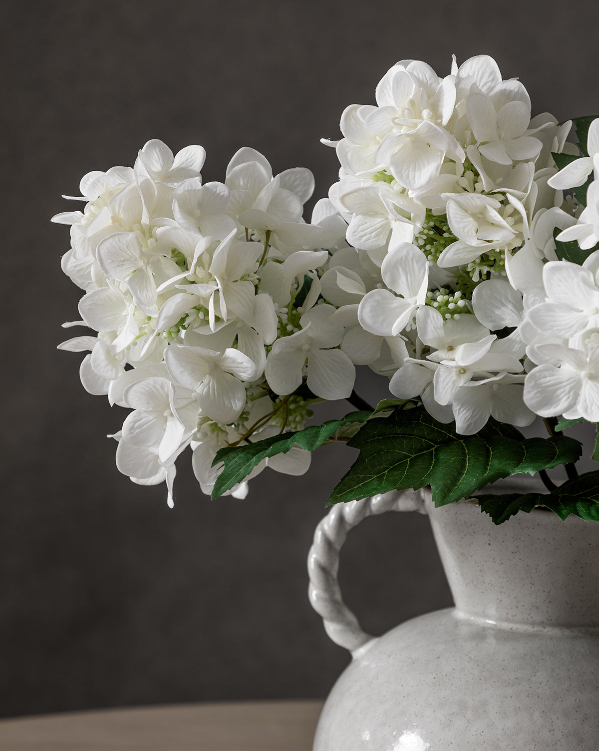 A close-up of a Faux Hydrangea Stem arrangement in a textured white ceramic vase with a twisted handle, displayed against a plain dark background.