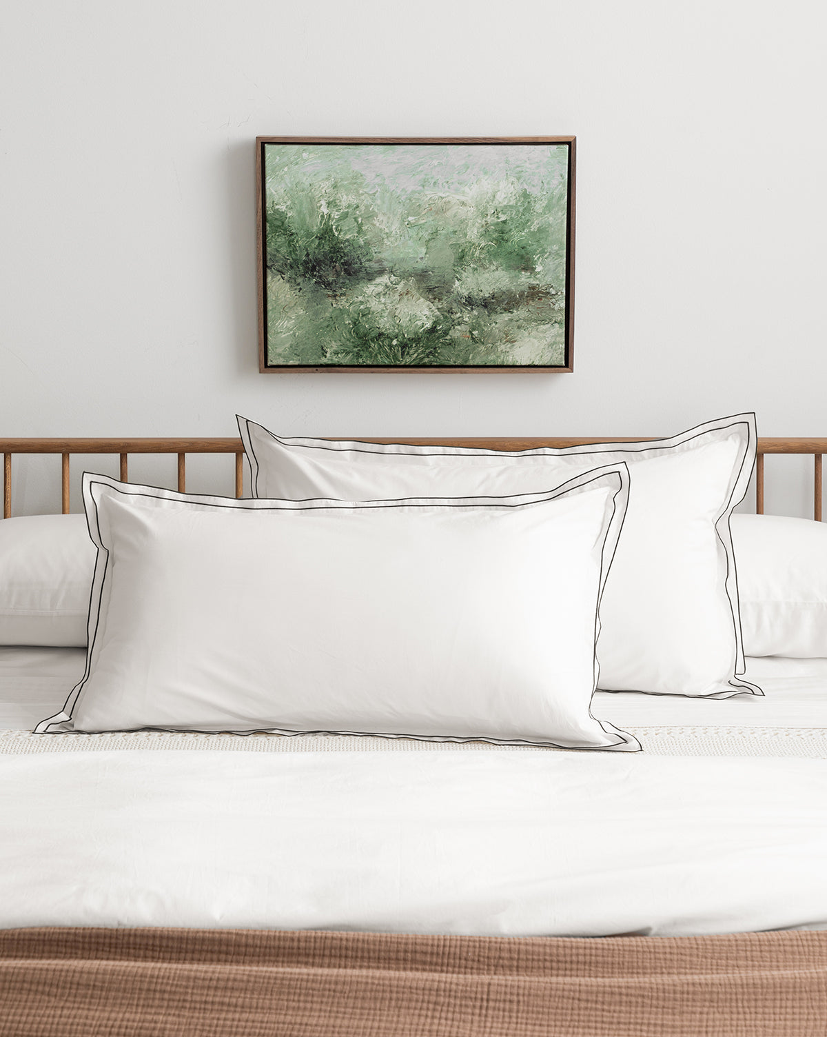 A neatly made bed with crisp white pillows featuring black piping, a wooden headboard, white bedding, a textured brown blanket at the foot, and Morning Cottage by Dan Hobday displayed as landscape art above the bed.