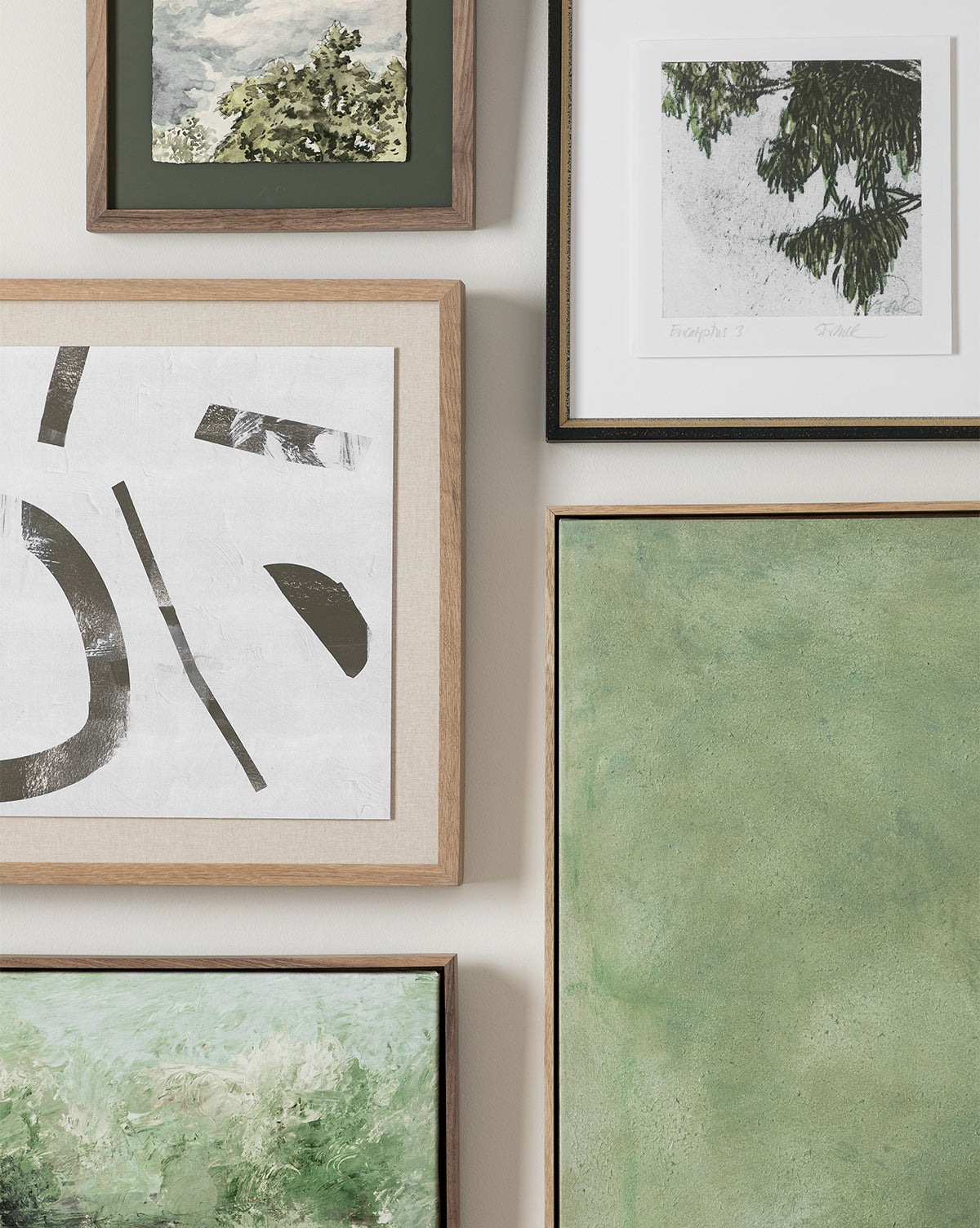 A gallery wall featuring Moss—a giclee abstract black and white print and green-toned landscape in natural wood frames—closely arranged on a light-colored wall.