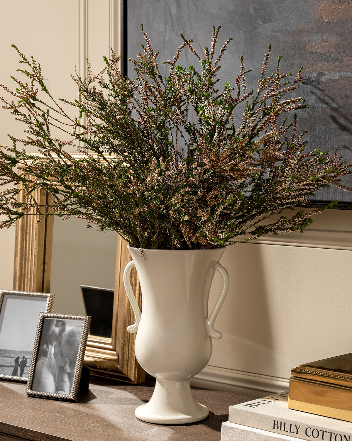 The Delphine Footed Vase, a tall and elegant white piece with handles, displays greenery on a wooden surface beside framed photos, books, and a gold-bordered mirror, with part of a painting visible in the background, by Vendixs