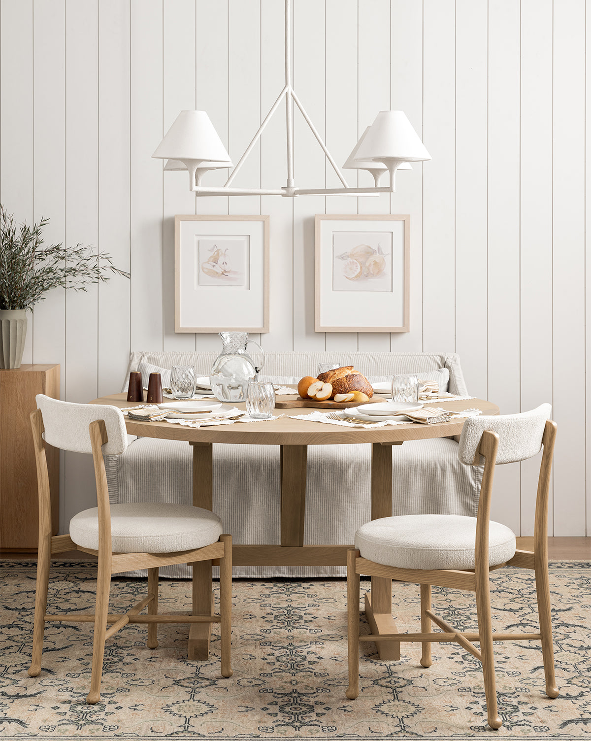 A cozy dining area with a wooden table set for breakfast, two cushioned chairs, and the Olivier Slipcover Dining Bench with feather and down cushions. Pastries, glassware, framed art, soft lighting, and white shiplap walls complete the inviting scene by McGee & Co.