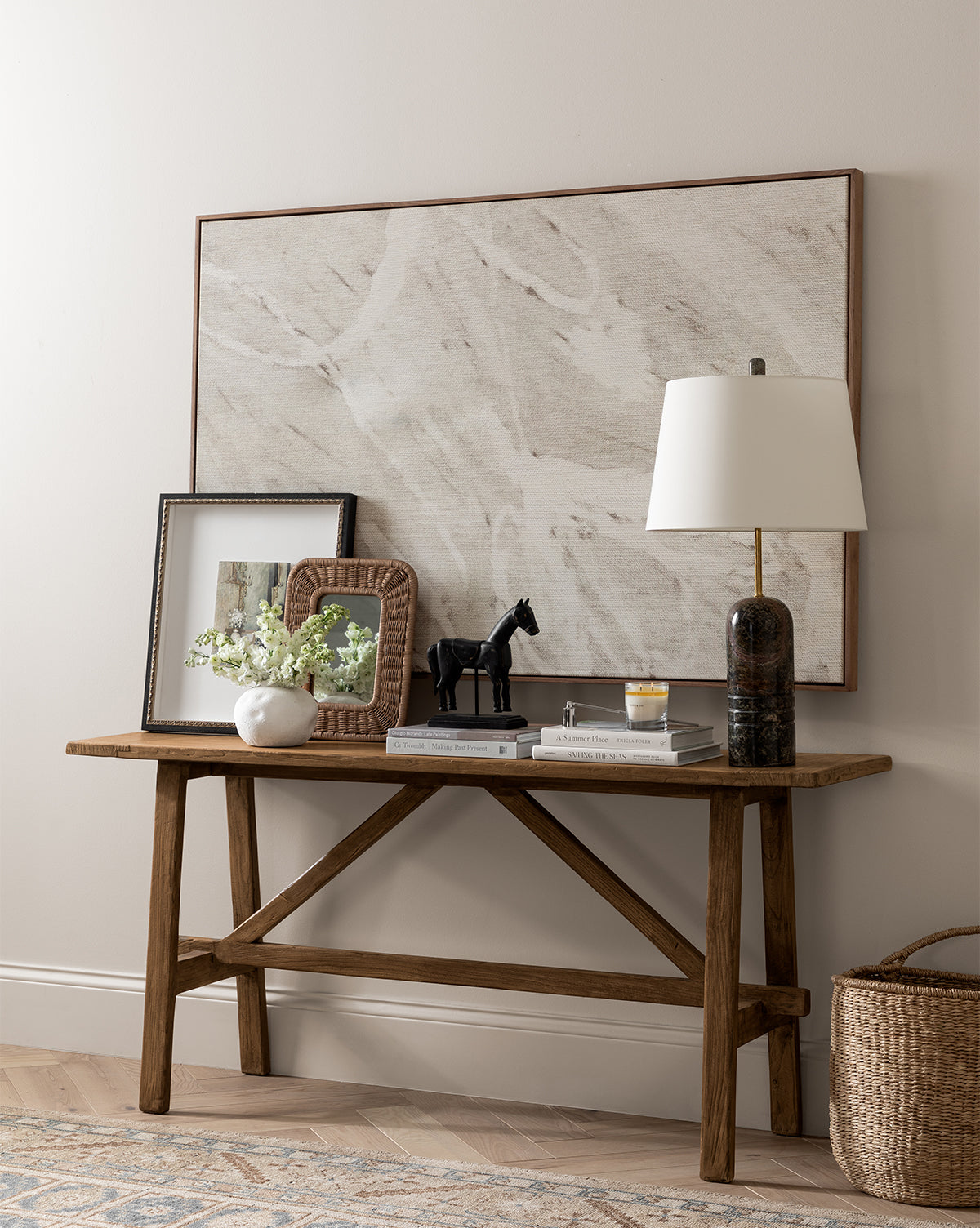 The Hampstead Console Table, crafted from reclaimed elm wood, displays a lamp, books, vases with flowers, framed art, a candle, a black horse figurine, and an abstract painting set against a beige wall by McGee & Co.