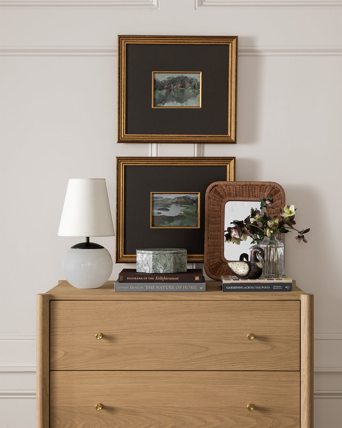 A light wood dresser topped with books, a marble box, a wicker-framed mirror, and a vase of flowers; the Tiny Terri Accent Lamp with its natural paper shade sits beside two framed paintings hanging on the wall above by Vendixs