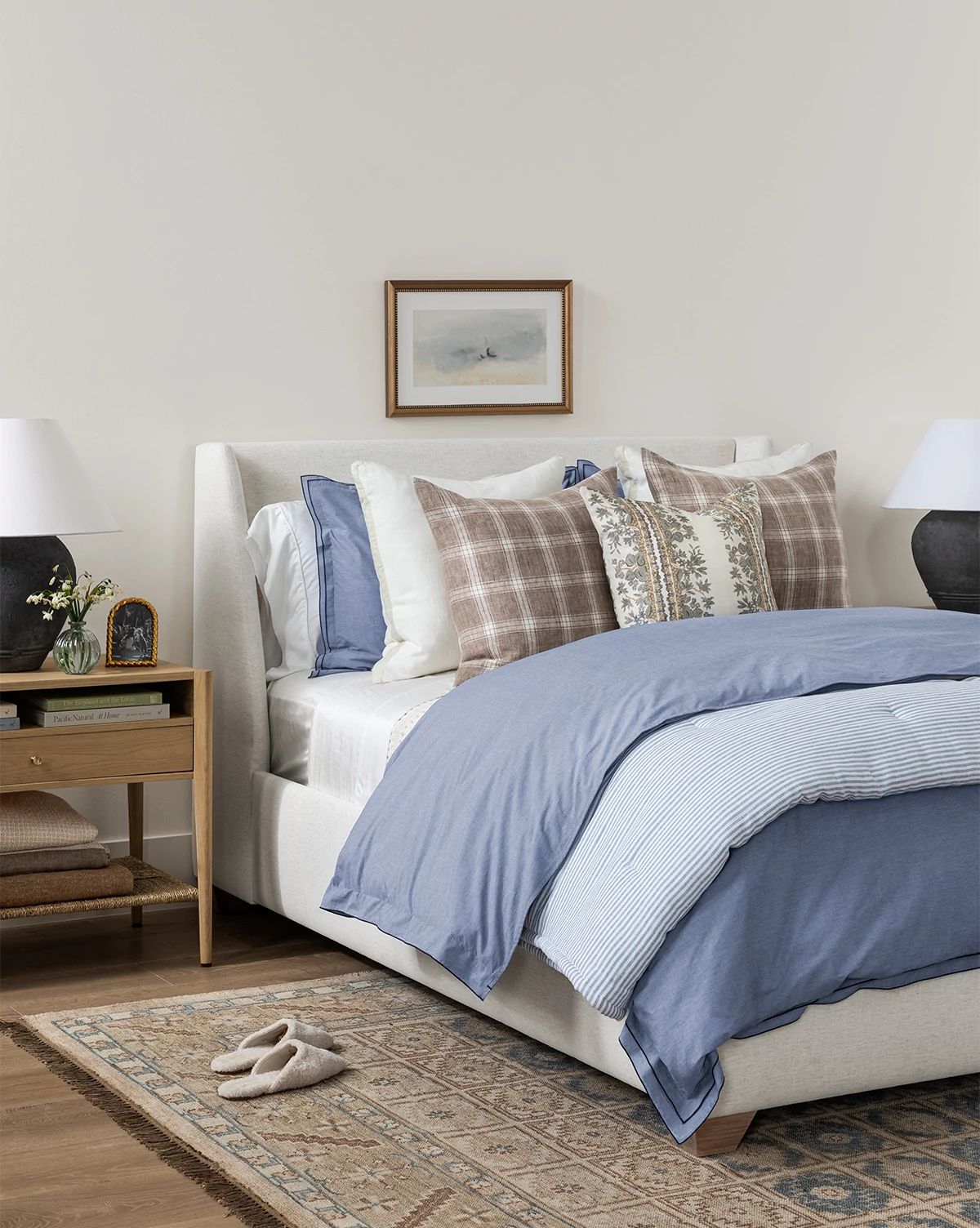 A cozy bedroom showcases the Community Walt Bed Low Headboard with blue bedding, decorative pillows, a nightstand with lamp, books, flowers, framed art above the bed, and slippers on a patterned rug beside its solid oak legs by Vendixs