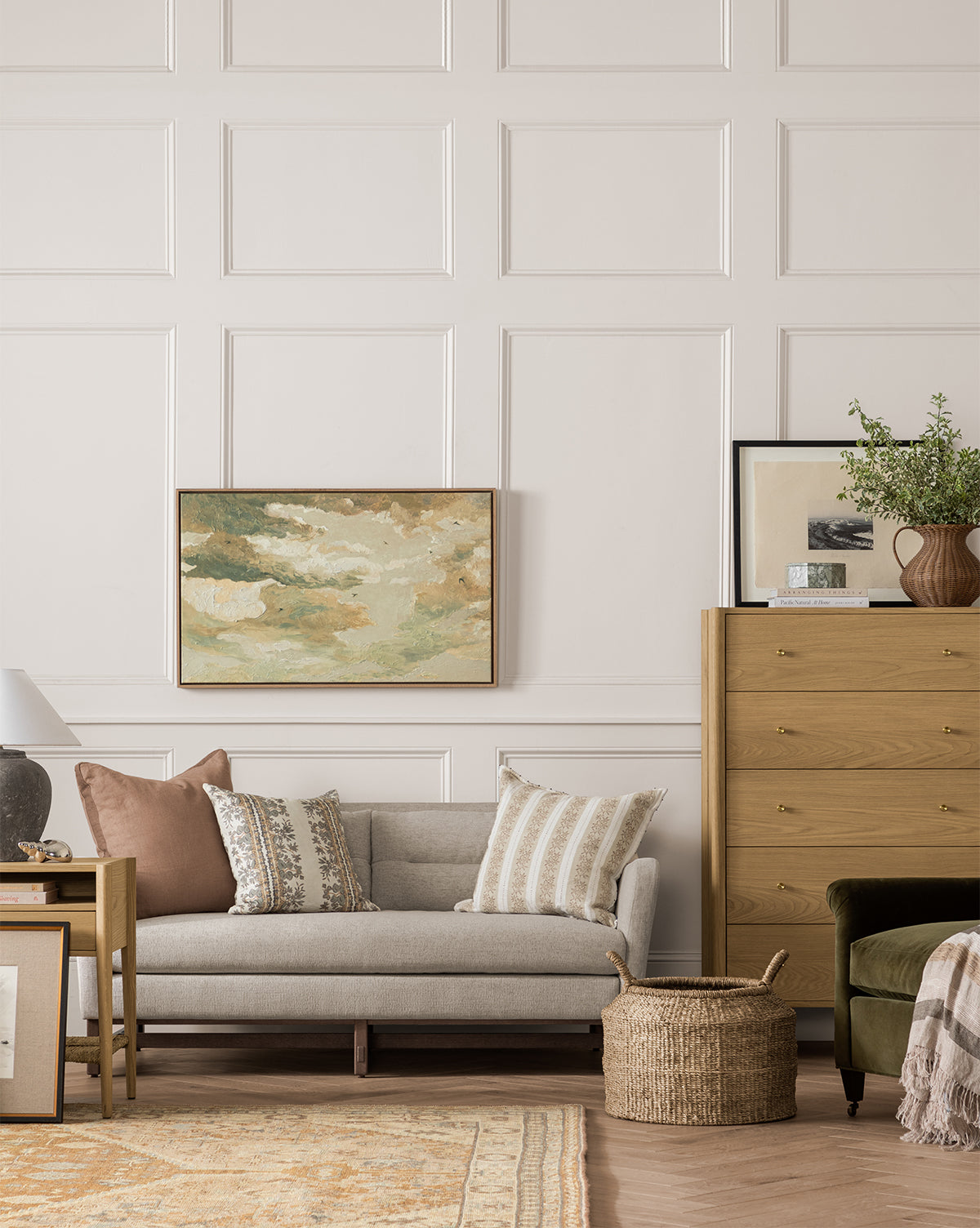 A cozy living room with a light gray sofa, patterned pillows, wooden side table, basket, tall dresser with decor, and the Swallows at Dusk landscape canvas in a white oak frame on neutral paneled walls by Vendixs