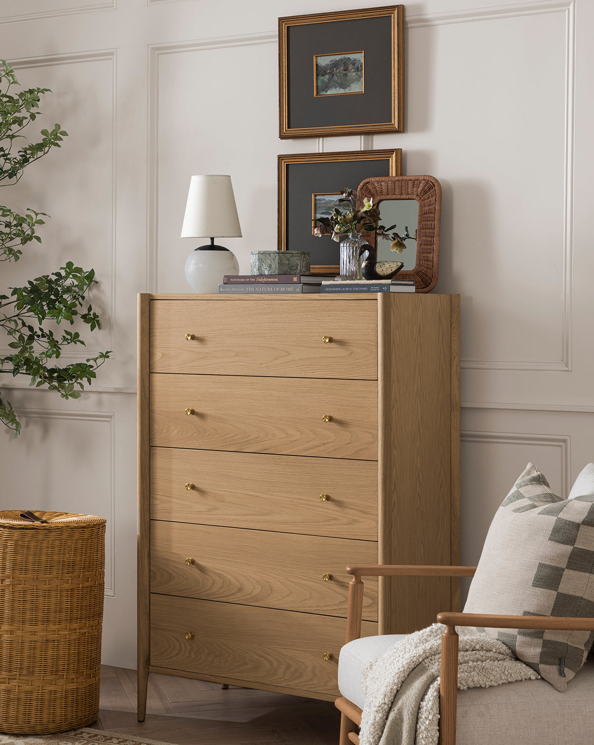 The Dana Tallboy Dresser with brass hardware stands against a white paneled wall, topped with a lamp, books, plant, and mirror. Framed art hangs above, while a chair and woven basket sit nearby with a leafy plant on the left by McGee & Co.