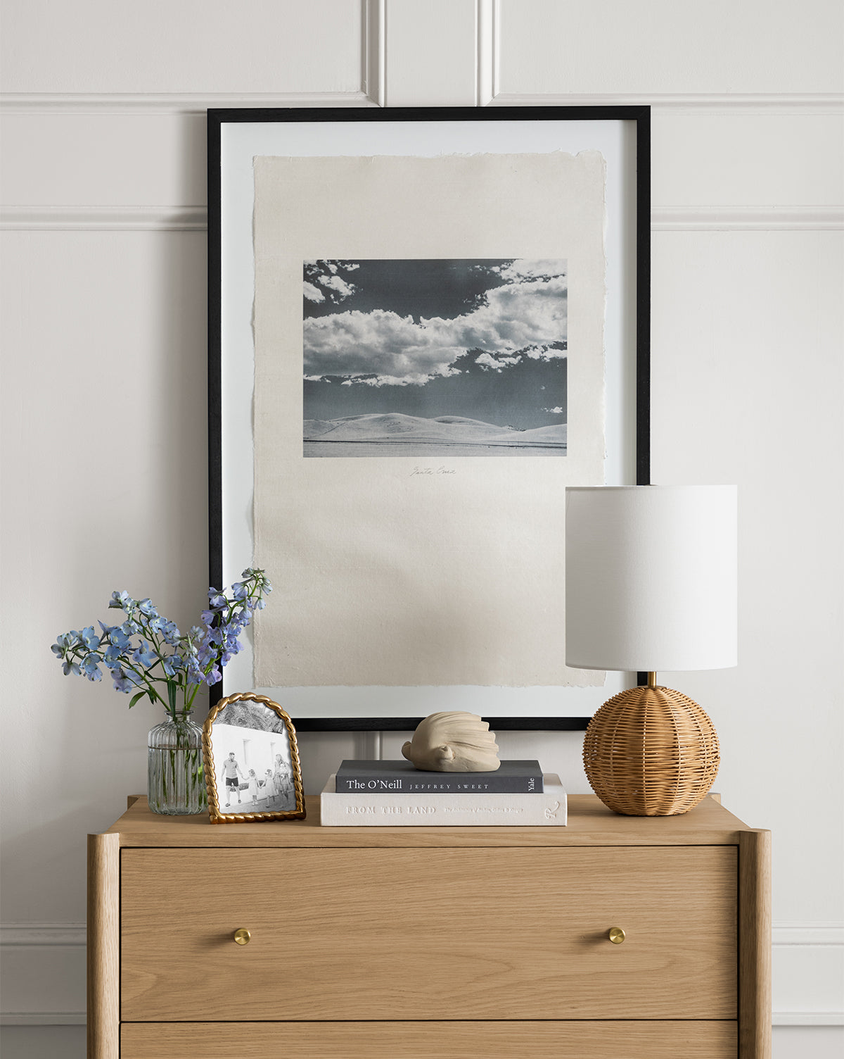 A wooden dresser with gold knobs displays a Mari Table Lamp with a white linen shade and rattan base, blue flowers in a vase, a framed photo, sculpture, and two books. Above is a framed black-and-white landscape photo by Vendixs
