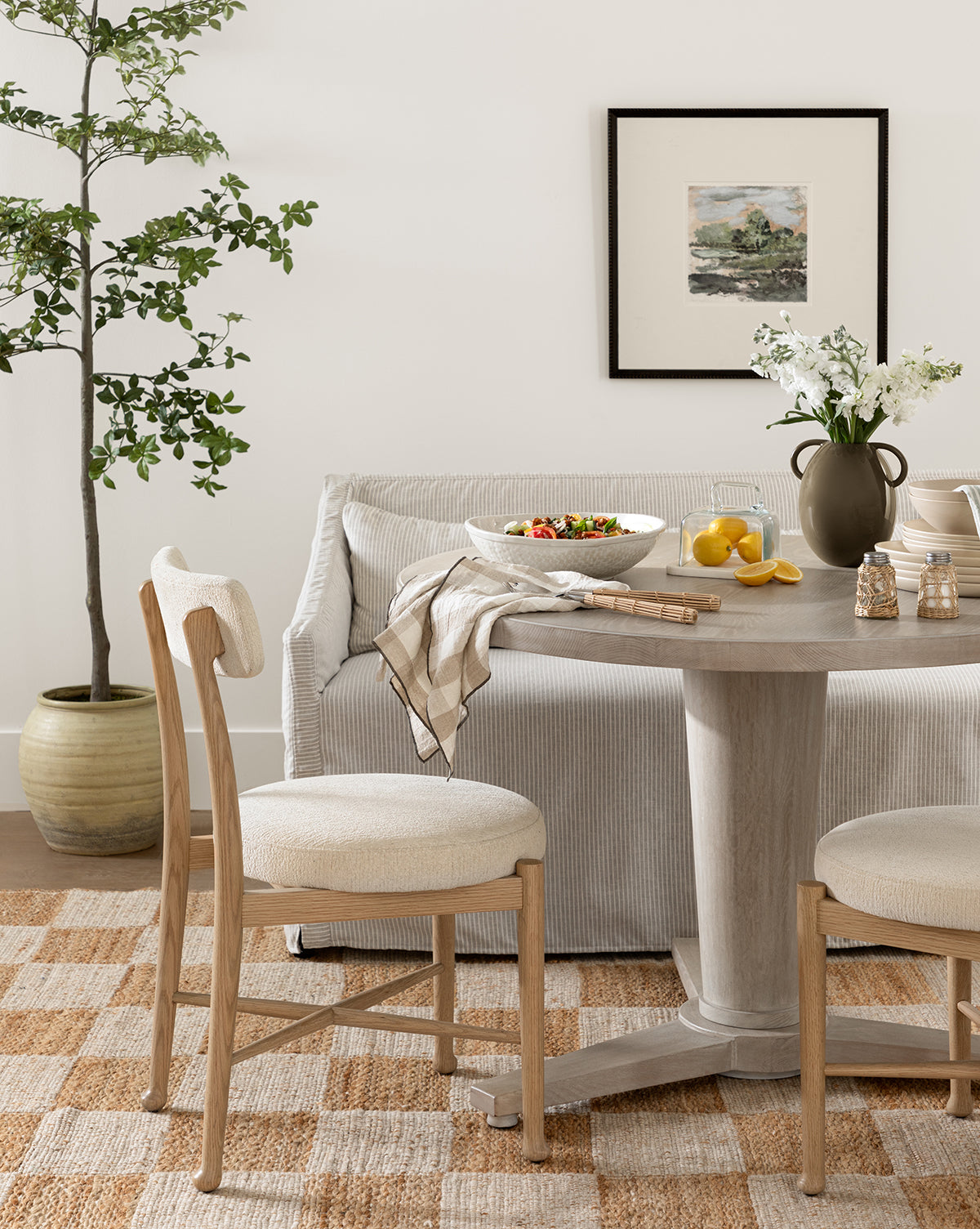 A cozy dining area features a round wooden table set for a meal, two cushioned chairs, a striped sofa, the Olivier Slipcover Dining Bench with feather and down cushion, a potted plant, framed artwork, and a checkered rug by McGee & Co.