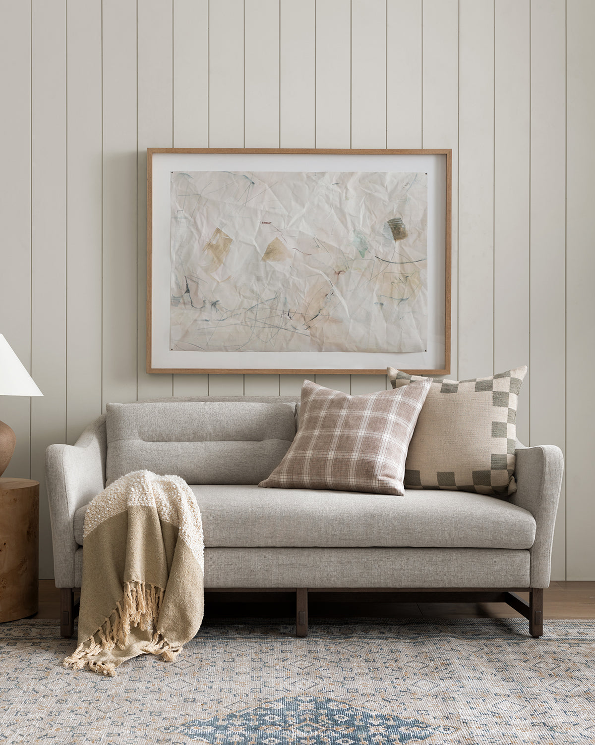 A neutral-toned living room features a light gray sofa with two patterned pillows, a beige throw blanket, abstract artwork on a paneled wall, and the Merilyn Hand-Knotted Rug adding warmth underfoot, by Vendixs