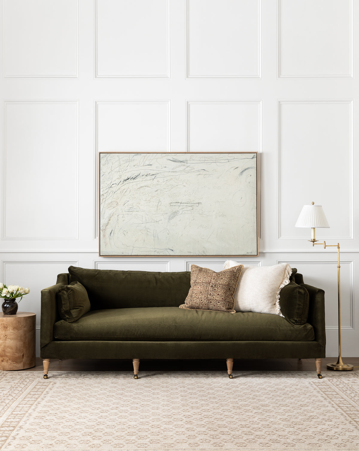 A living room featuring the Rowe Fine Furniture Haverford Upholstered Sofa 90 in olive green velvet with three assorted pillows, a round wooden side table with flowers, a floor lamp, and an abstract painting above a light patterned rug, blending seamlessly with Vendixs