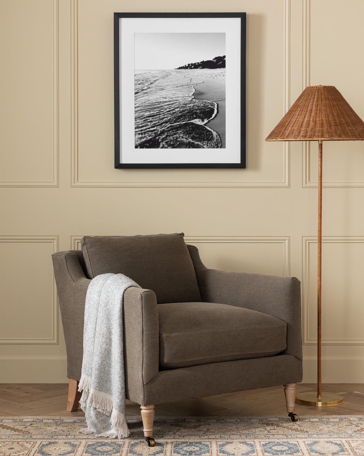 The Haverford Upholstered Lounge Chair by Rowe Fine Furniture, Inc. in brown, with a light gray throw, sits beside a wicker floor lamp. Above, a black-and-white beach photo hangs on beige paneling over a patterned rug. Vendixs