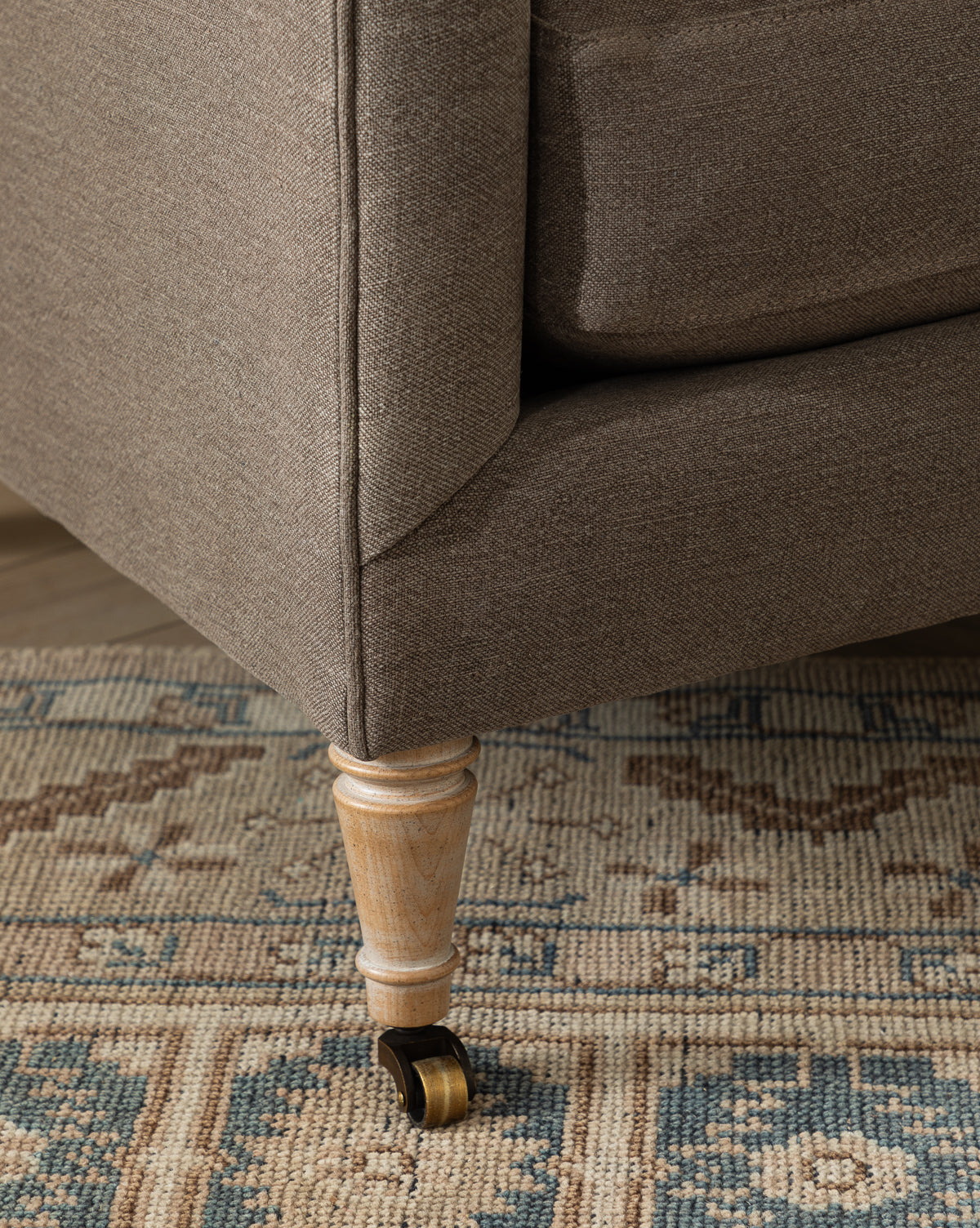 Close-up of the Haverford Upholstered Lounge Chair, featuring brown upholstery and a light wooden leg with a caster wheel, set on a patterned beige and blue rug, by McGee & Co.