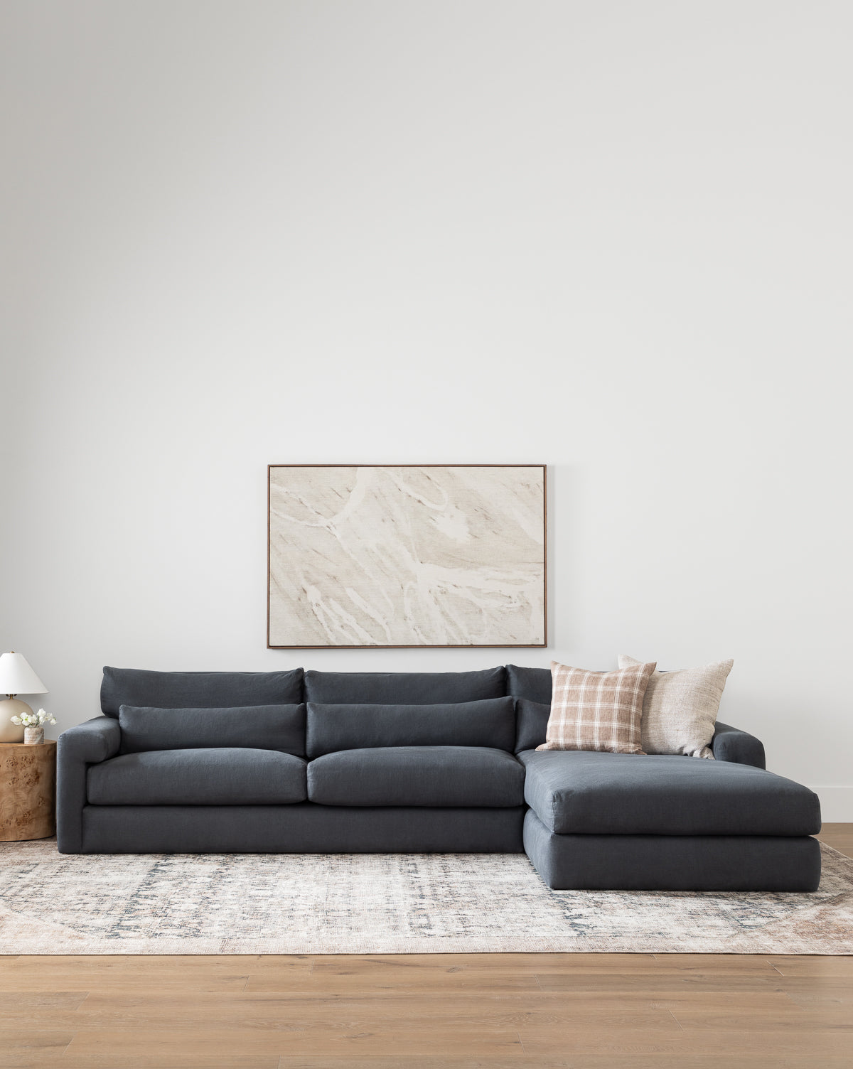 A modern living room with the Community Beckstead Chaise Sectional in dark grey, a light patterned rug, a round wooden side table with a lamp, and neutral abstract art on a white wall. Vendixs