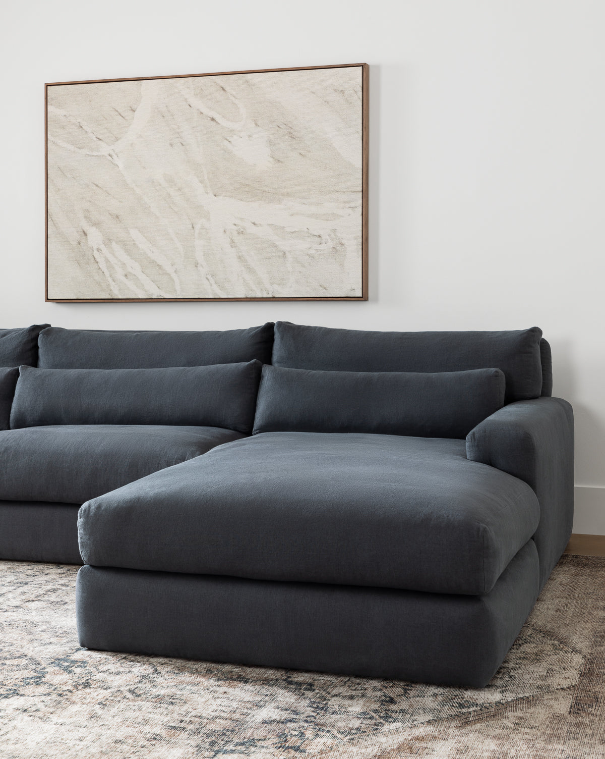 A modern living room featuring the Community Beckstead Chaise Sectional in dark gray, a textured area rug, and a large neutral-toned abstract artwork displayed on the white wall above the sofa, Vendixs