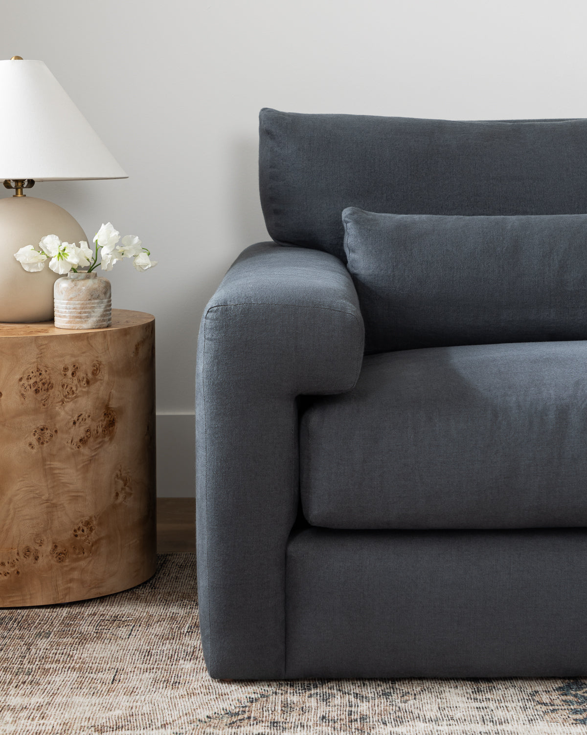 A modern living room featuring the Community Beckstead Chaise Sectional in dark gray, paired with a round wooden side table, a beige lamp, and a small vase of white flowers atop a neutral patterned rug, finished with touches from Vendixs