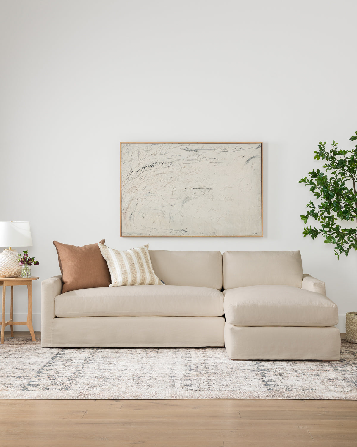 A modern living room features the Community Macy Slipcover Chaise Sectional 101, two throw pillows, a patterned rug, a wooden side table with a lamp and decor, a large abstract painting, and a leafy green plant in the corner—all styled with pieces from Vendixs