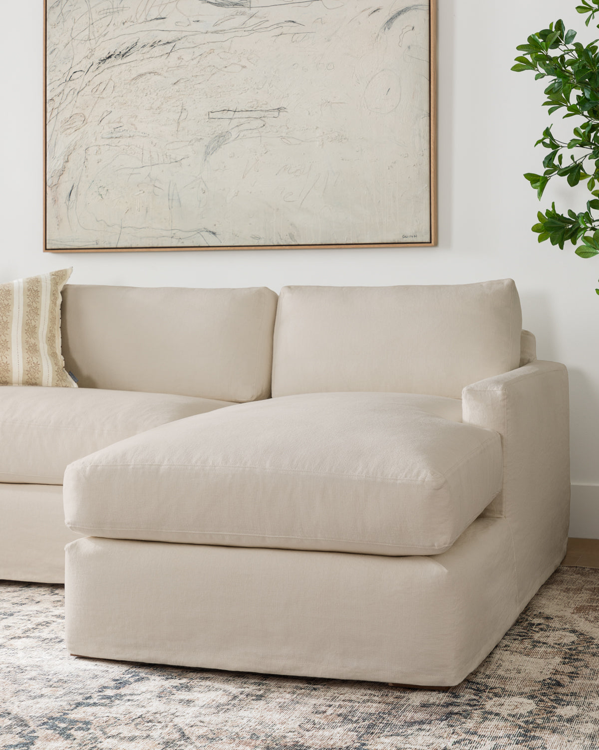 A modern living room features the Community Macy Slipcover Chaise Sectional 101 in beige, paired with a patterned throw pillow, an abstract wall painting, a green plant, and a neutral-toned area rug from Vendixs