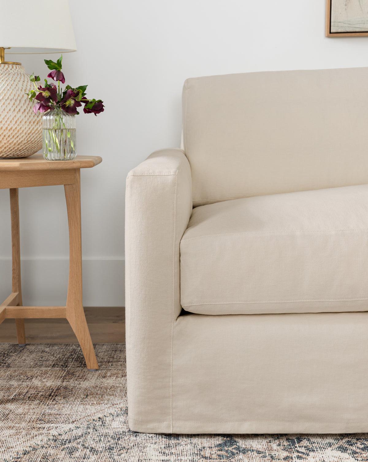 A beige upholstered armchair sits by a wooden side table with a glass vase of purple flowers and a white lamp, on a neutral patterned rug—perfectly complementing the Community Macy Slipcover Chaise Sectional 101 by Vendixs