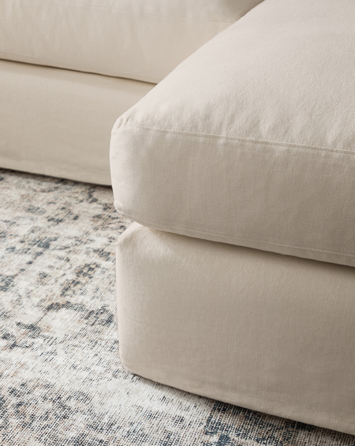 A close-up of the Community Macy Slipcover Chaise Sectional 101 in beige upholstery with large, plush cushions, set on a textured, patterned neutral-toned area rug, Vendixs