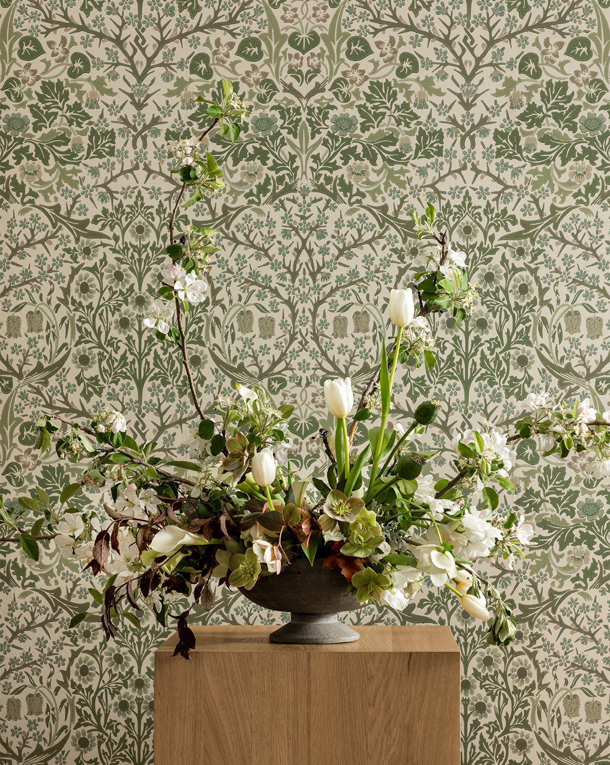 A large floral arrangement with white tulips and greenery in a gray vase sits on a wooden pedestal, set against Morris & Co. x Vendixs Blackthorn Wallpaper by Sanderson Design Group, Inc., featuring a botanical green and cream design by Vendixs