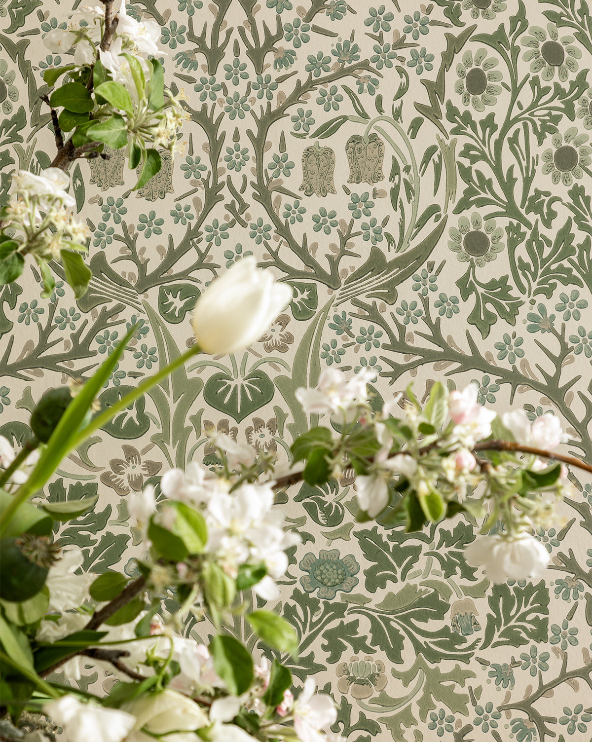 Flowering branches with white blossoms and green leaves stand before Sanderson Design Group, Inc.s Morris & Co. x Vendixs Blackthorn Wallpaper, which features an intricate floral pattern in muted green and beige tones, Vendixs