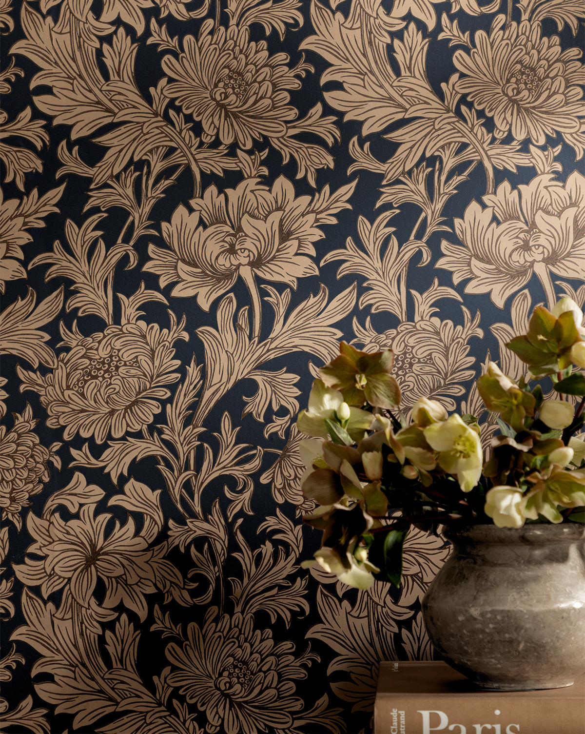 The Morris & Co. x Vendixs Chrysanthemum Wallpaper by Sanderson Design Group, Inc. features bold beige floral motifs on a dark background, evoking classic style. A vase with pale flowers rests on books in the lower right corner of the scene—McGee & Co.