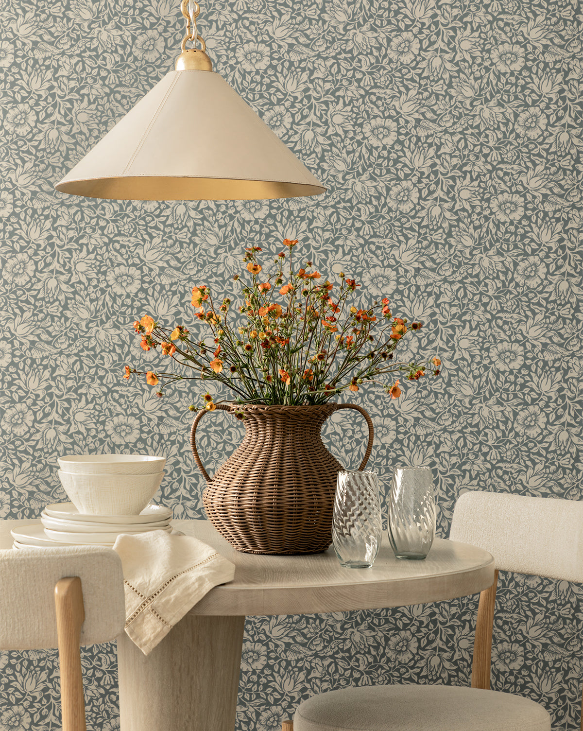 A round wooden table set with white dishes and textured glasses sits under a cream pendant lamp. A wicker vase with orange wildflowers is centered, backed by Morris & Co. x Vendixs Mallow Wallpaper by Sanderson Design Group, Inc., blending timeless charm with the signature style of Vendixs