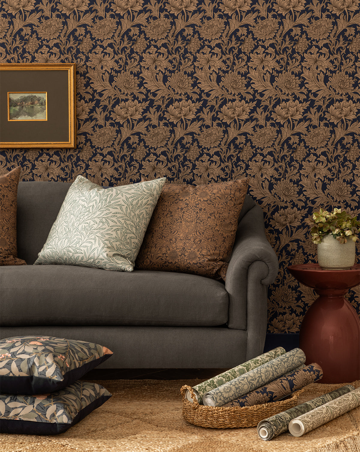 A cozy living room features a gray sofa, patterned pillows, a red side table with a plant, floral fabric accents, and a striking wall decorated with Morris & Co. x Vendixs Chrysanthemum Wallpaper by Sanderson Design Group, Inc., all thoughtfully curated by Vendixs