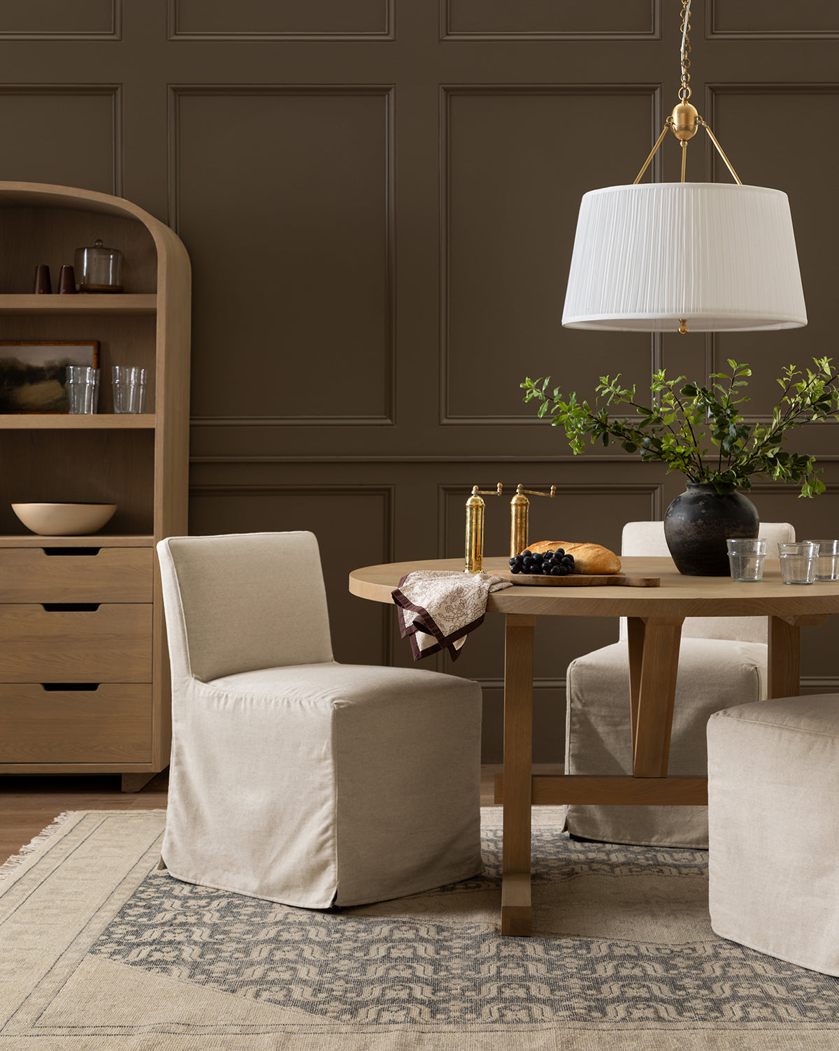 A modern dining room with neutral tones features a round wooden table, Olivier Slipcover Dining Chairs, a large pendant lamp, a patterned rug, a shelf sideboard, and a vase with green branches as the centerpiece, by Vendixs