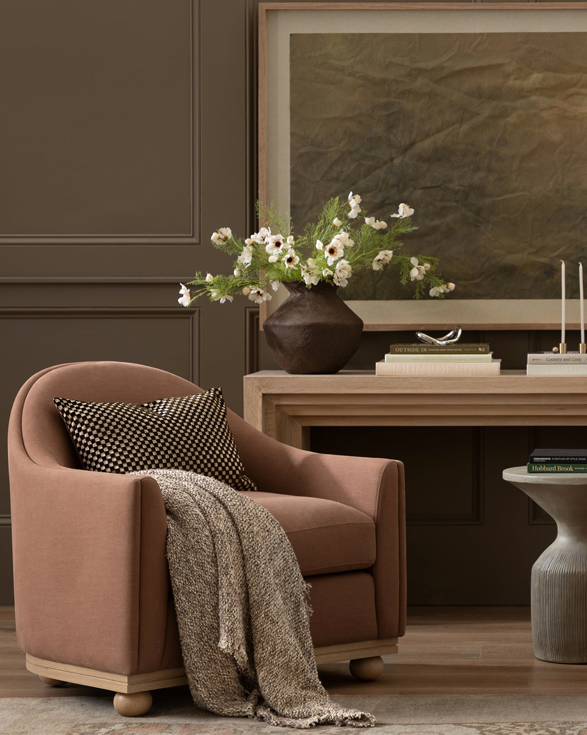 A mauve armchair with a throw and cushion sits beside a wooden desk. On the desk is a large vase of Cream Cosmos Stem, candles, and abstract art, all set against a brown paneled wall by McGee & Co.