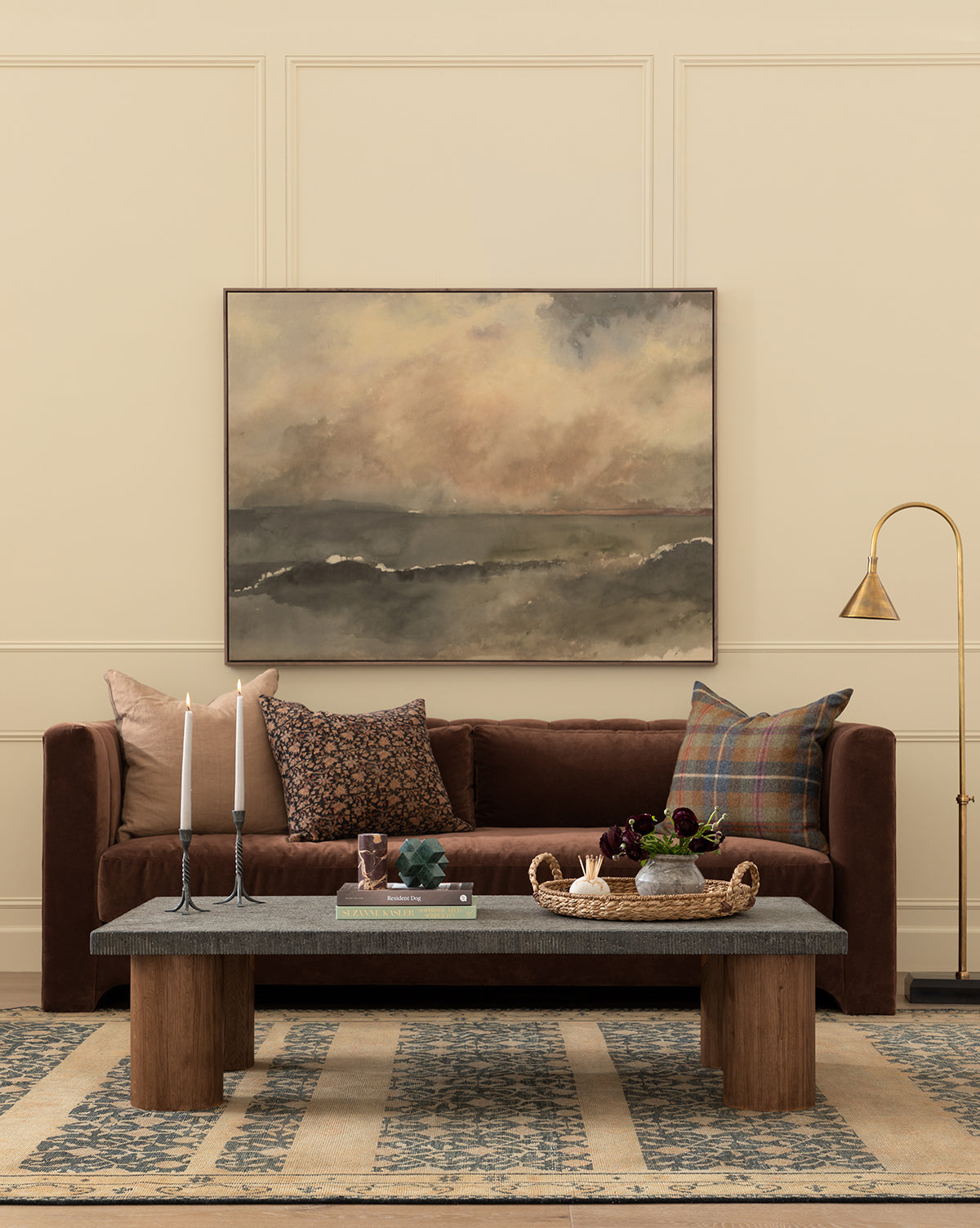 A cozy living room features a brown sofa with patterned pillows, a wooden coffee table with decor, tall candles, the gallery-quality You Said I am the Sea by Aileen Fitzgerald painting, a brass floor lamp, and a blue patterned rug on beige flooring, by Vendixs