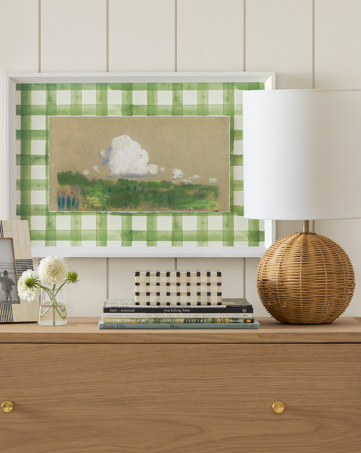 A wooden dresser holds stacked books, a photo frame, and a round wicker lamp with white shade. Above hangs a landscape painting with a green checkered border, echoing the style of Oliver Jeffers’ This Moose Belongs to Me.