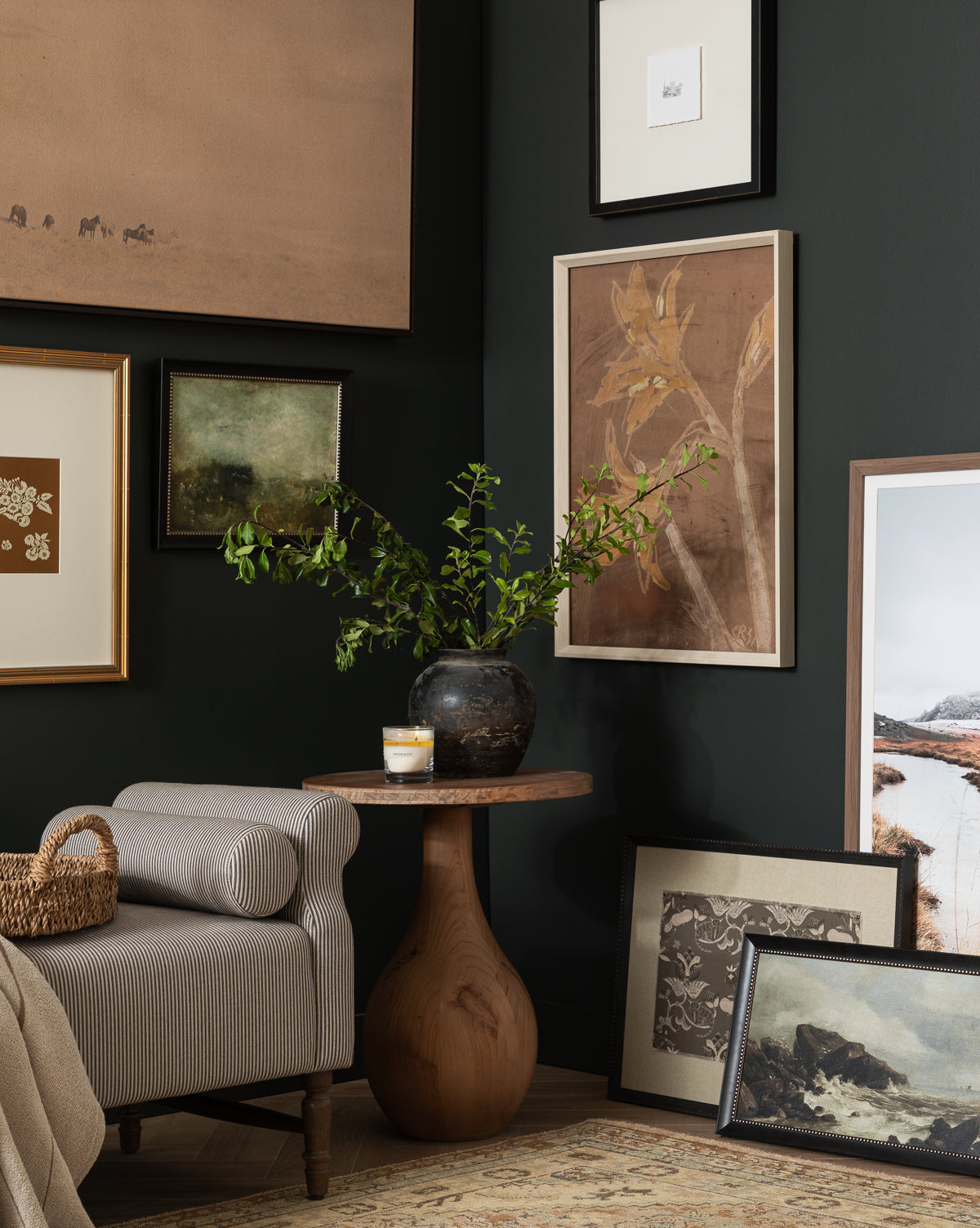 A cozy living room corner features a striped armchair and a round wooden table with a dark vase, candle, and green branches. Past Petals giclee print art hangs on dark walls, with additional white wood frames leaning on the floor, by Vendixs