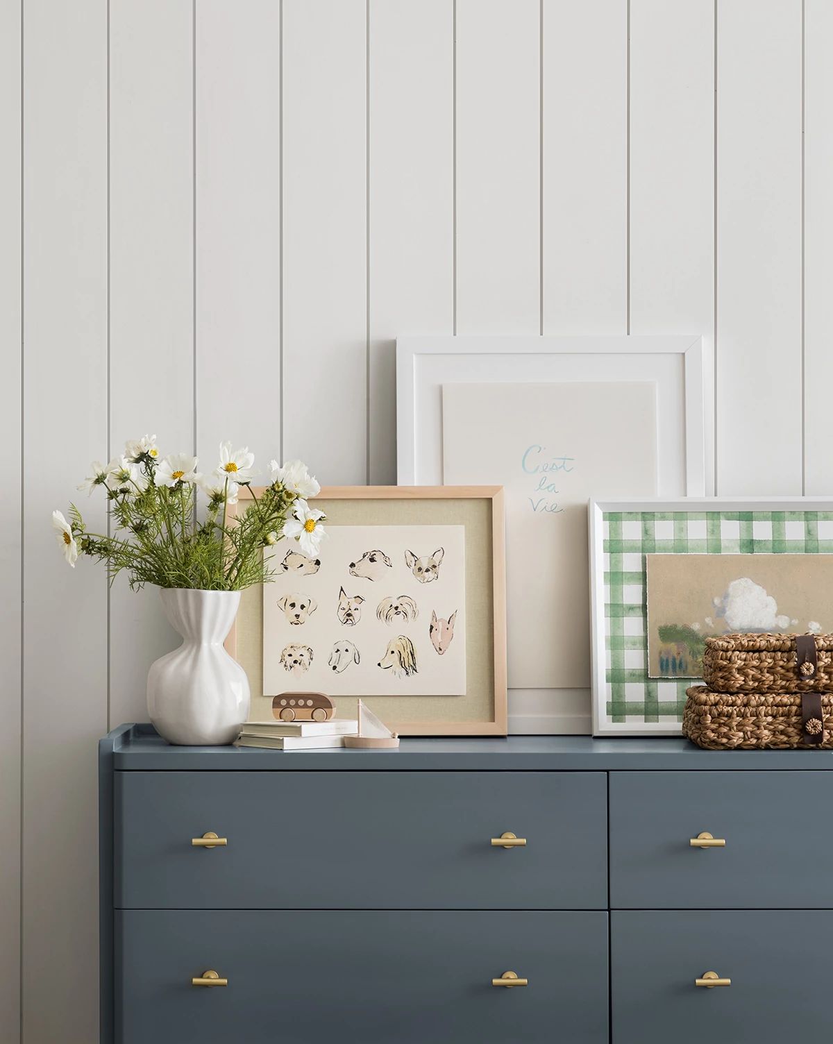 A blue dresser with gold handles displays a white vase of daisies, framed art, woven baskets, and the Woof ceramic dog dish against a white shiplap wall, by Vendixs