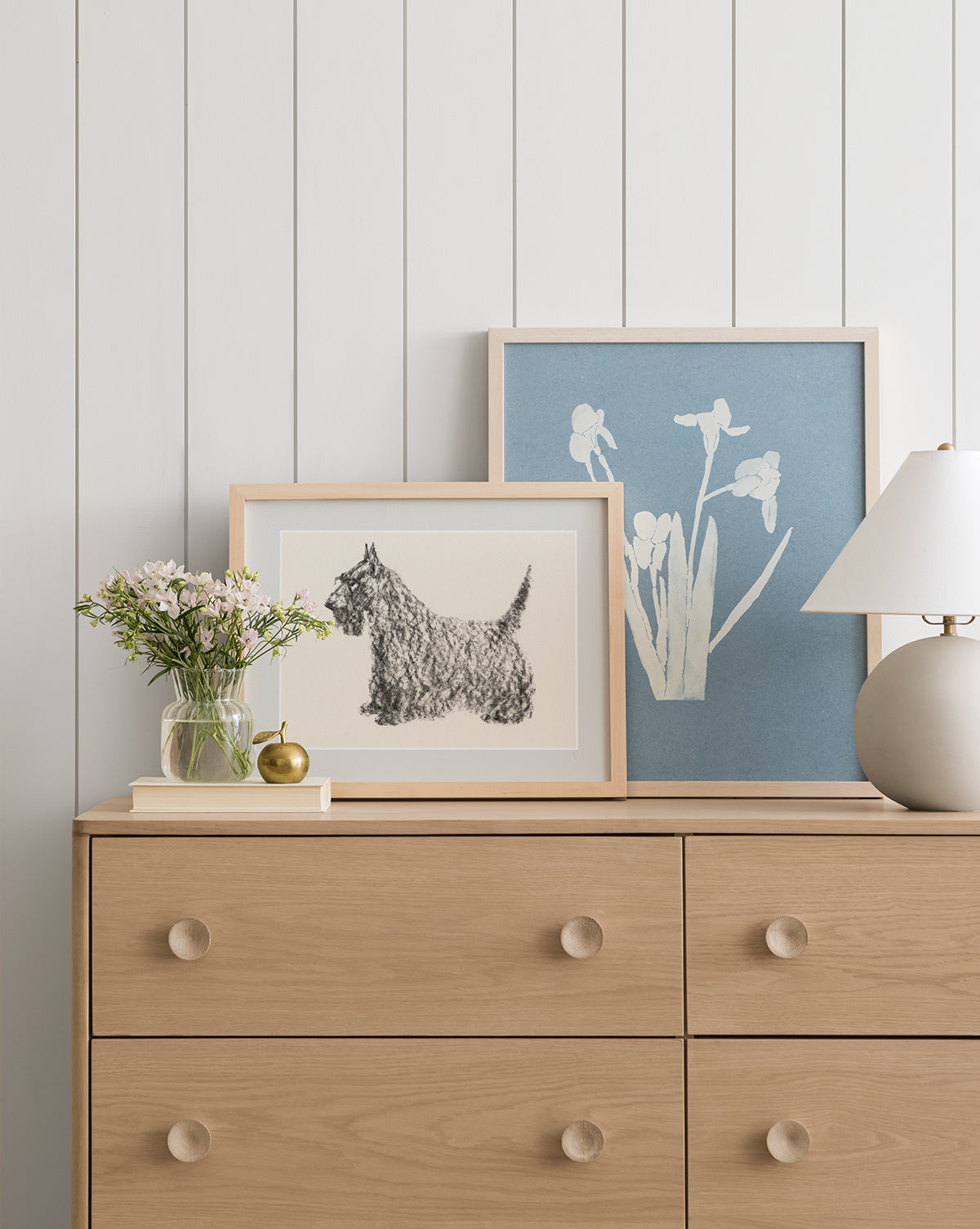 The Finn light wood dresser with round knobs holds a small glass vase of flowers, a gold trinket, and a white table lamp. Two framed artworks—a Scottish Terrier sketch and white blooms on blue—lean against a white paneled wall, by Vendixs
