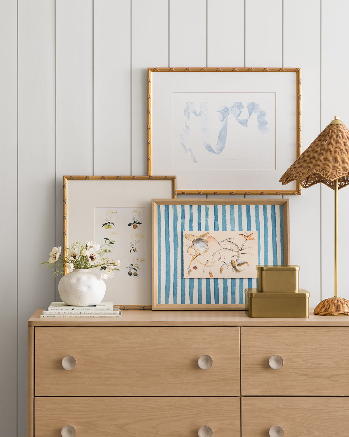 A light wood dresser with round knobs holds decor and three framed art prints—including the Loose Ribbons giclee print featuring blue stripes—against a white paneled wall in this cozy children’s room, by Vendixs
