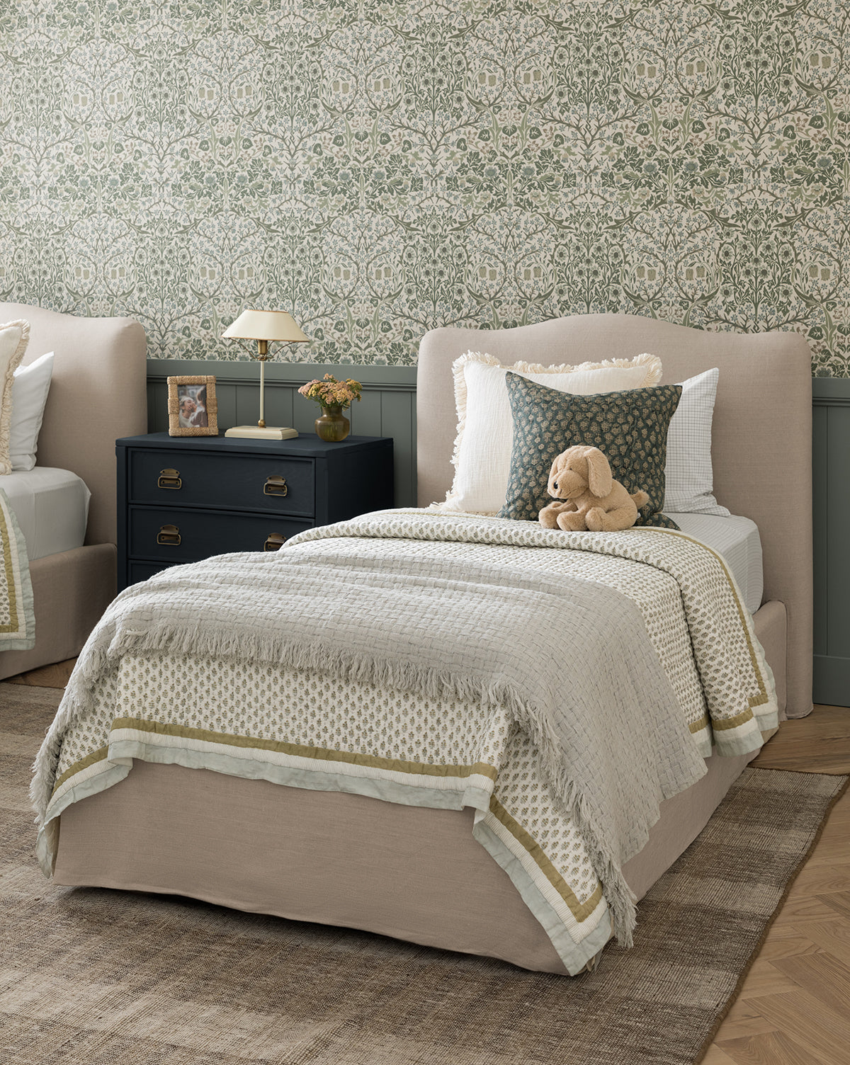 A neatly made bed with beige and white bedding, a patterned pillow, and the soft Caroline Kids Blanket by Indigo Craft sits next to a black nightstand with a lamp and books, set against floral wallpaper in this cozy, elegant bedroom—finished with timeless touches from Vendixs