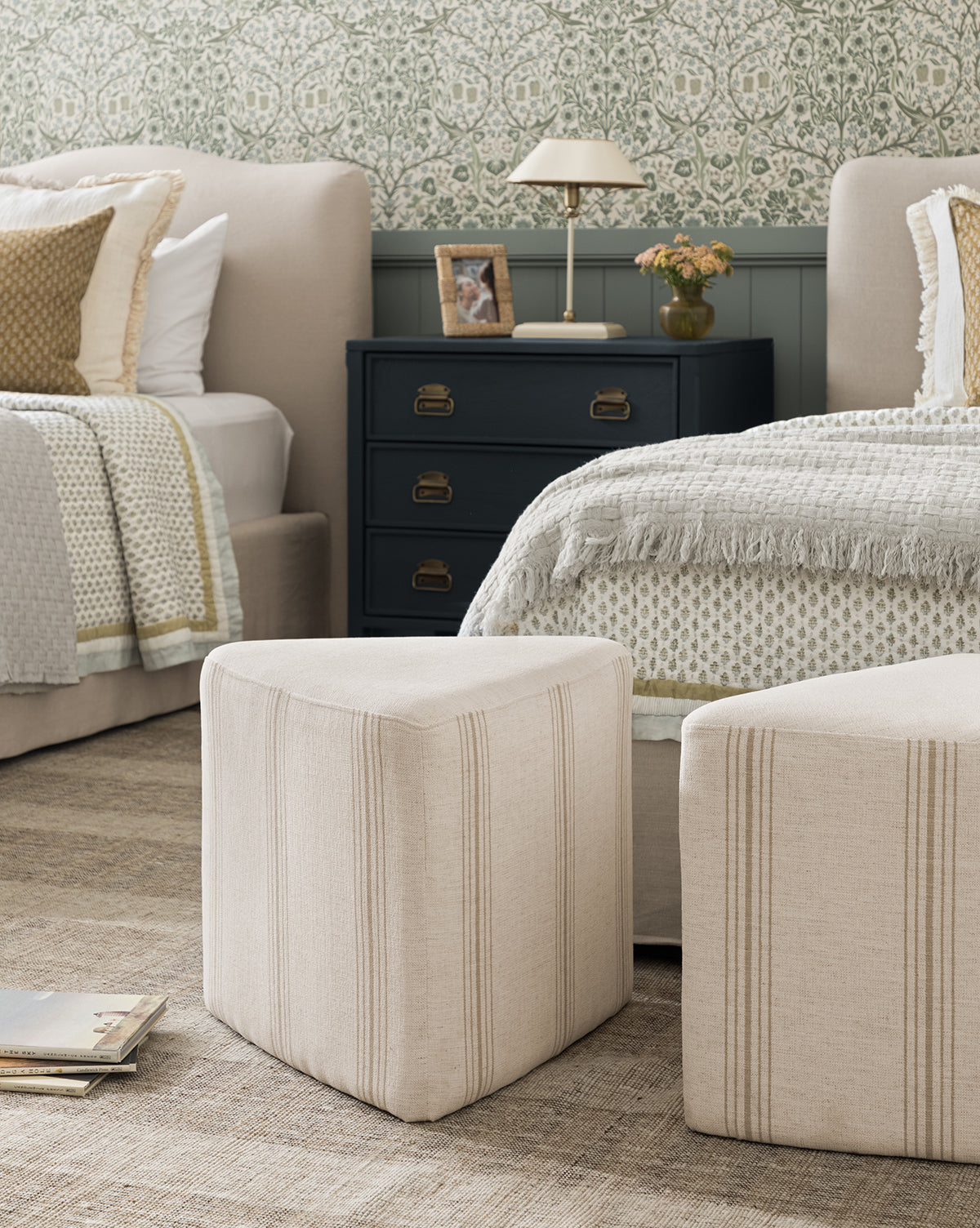 A cozy bedroom features two beige beds with textured bedding, a black nightstand with a lamp and flowers, two Jacques Ottomans by Dongguan City Baby Furniture Co., Ltd in crypton striped fabric, patterned wallpaper, and magazines on the woven rug—great for kids’ rooms, finished with accents from Vendixs