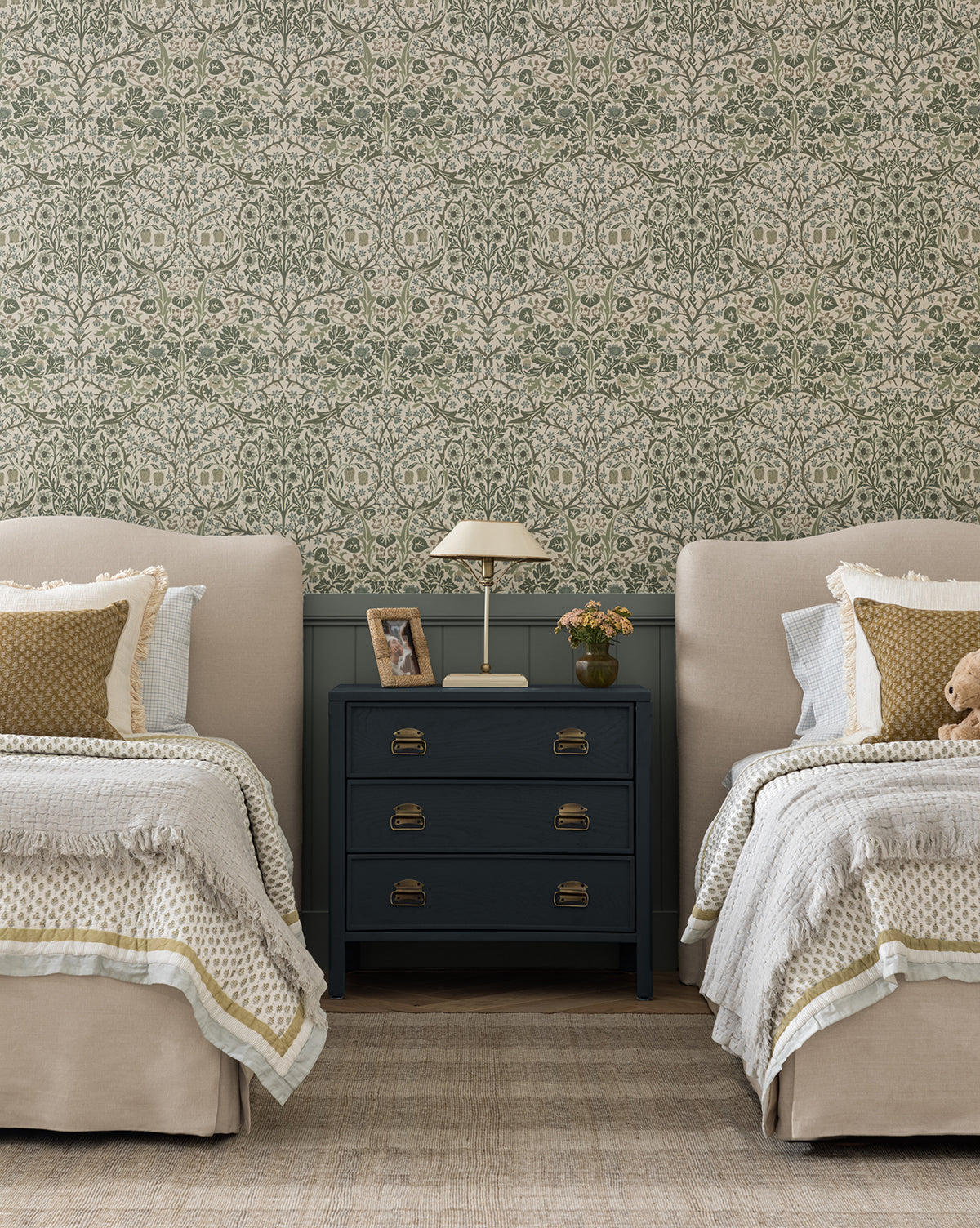 Two twin beds with beige headboards and bedding are separated by a navy nightstand with a lamp, photo, and book. Behind them, Sanderson Design Group’s Morris & Co. x Vendixs Blackthorn Wallpaper adds green floral charm; a teddy bear sits on one bed—a cozy space layered with thoughtful details from Vendixs