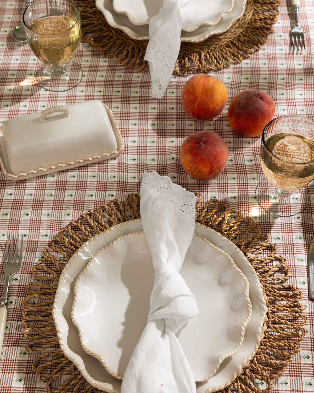 A table for two with scalloped white plates, woven placemats, wine glasses, a covered dish, three peaches, and Vendixs x Loeffler Randall Delilah Broderie Napkins (Set of 4) on a pink plaid Loeffler Randall tablecloth, by Vendixs