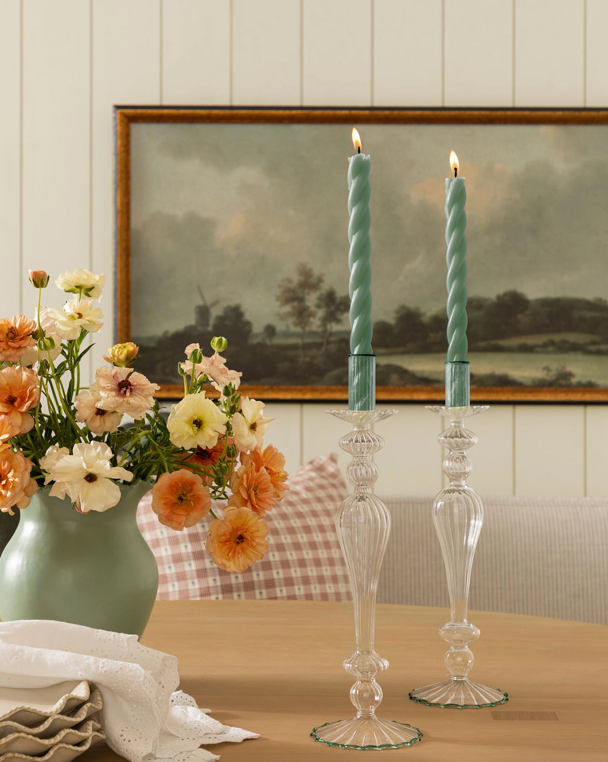 A vase of peach and cream flowers sits on a wooden table beside two lit, twisted blue candles in a Vendixs x Loeffler Randall Effie Taper Holder. A folded white napkin is nearby, with a landscape painting visible in the background by Vendixs