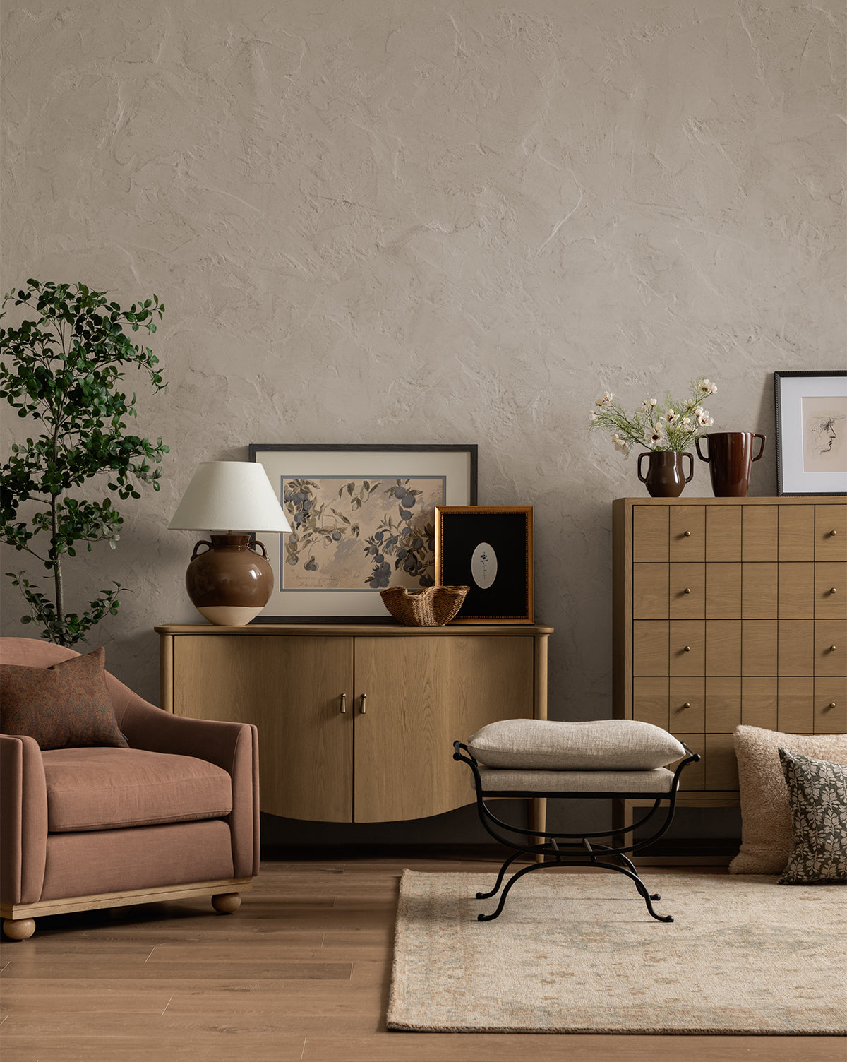 A cozy living room featuring Blue Fruits, pink armchair, beige ottoman, light wood cabinet, potted plant, decorative vases, botanical art in a charcoal frame, and a textured rug on wood flooring against a neutral wall, by Vendixs