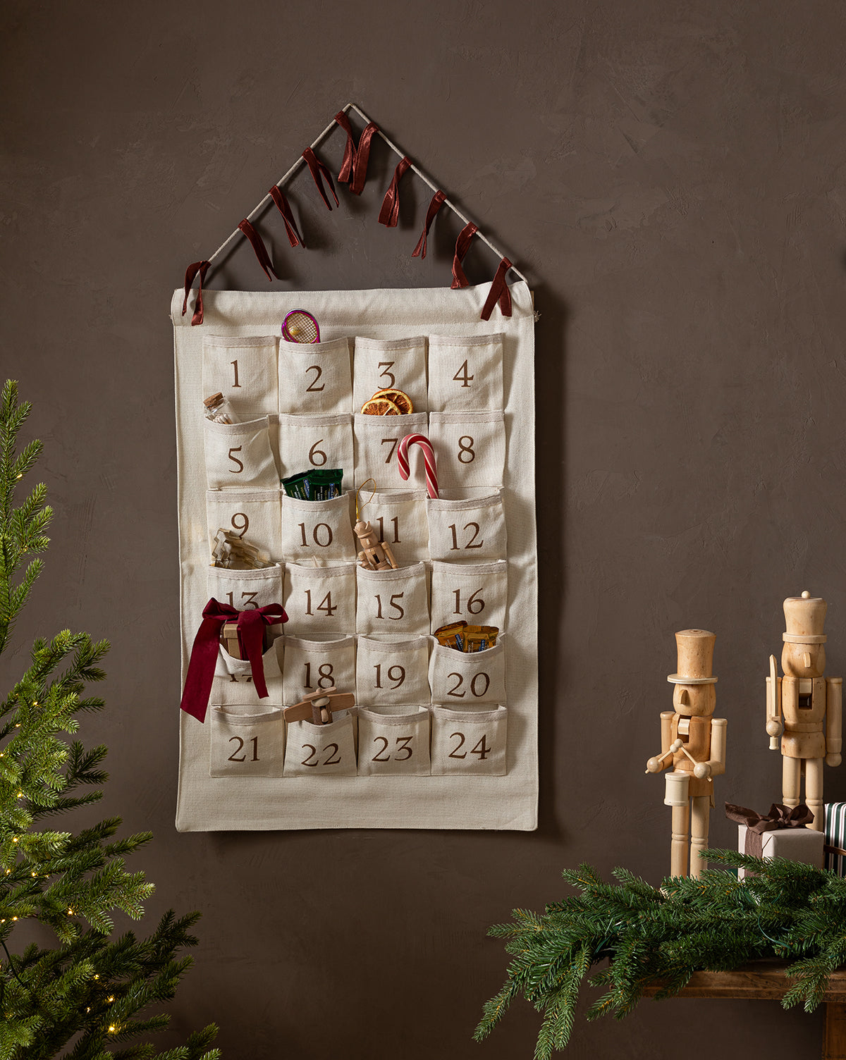 The Advent Calendar, made of cotton linen with numbered pockets for small gifts and ribbons, hangs on the wall. Below, two wooden nutcrackers and pine branches sit near a Christmas tree, creating a charming holiday scene, by Vendixs