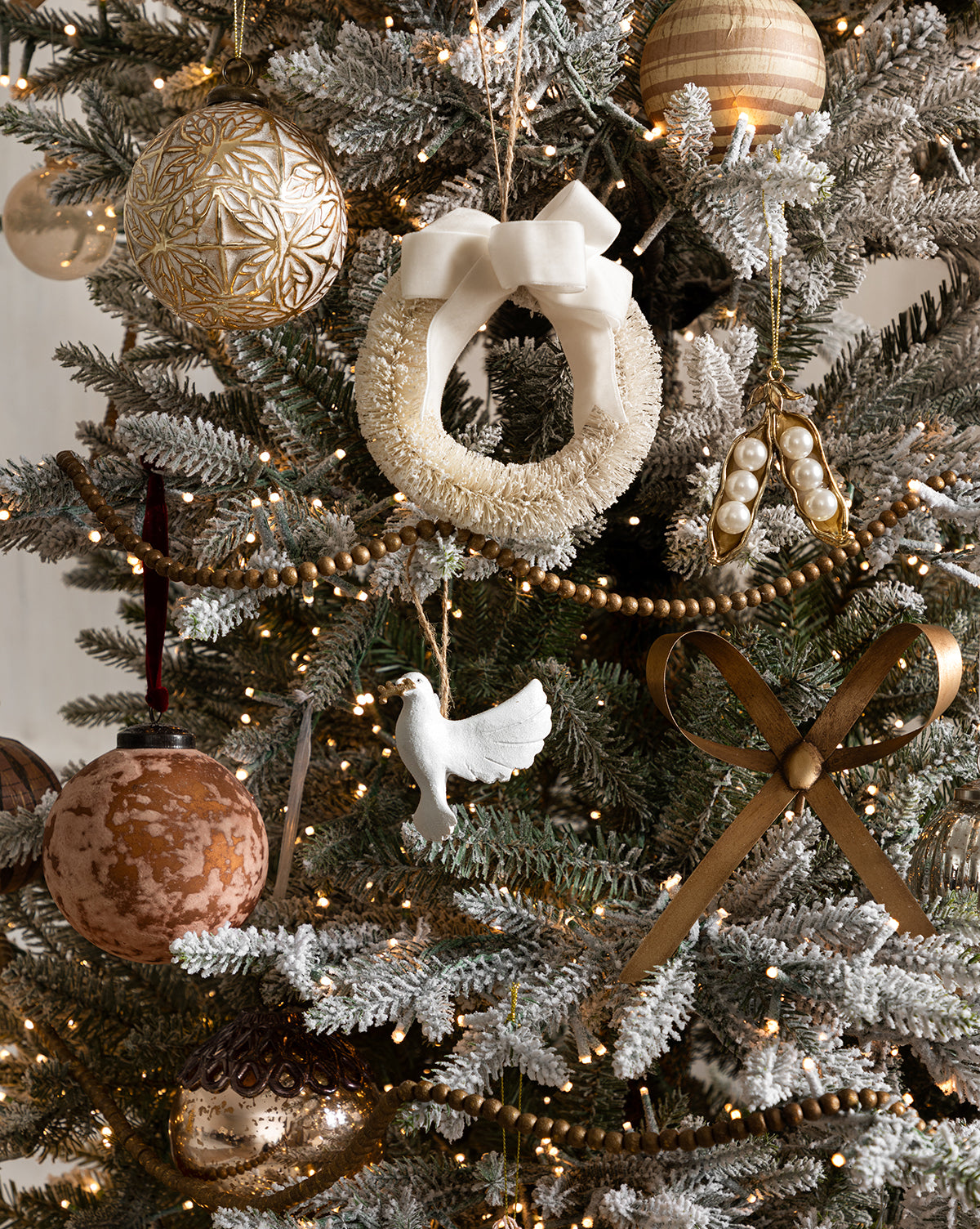 A Christmas tree decorated with gold and white ornaments features a wreath adorned with the Brass Bow Clip Ornament in antique brass, a white dove, gold baubles, beaded garland, and string lights, by Vendixs
